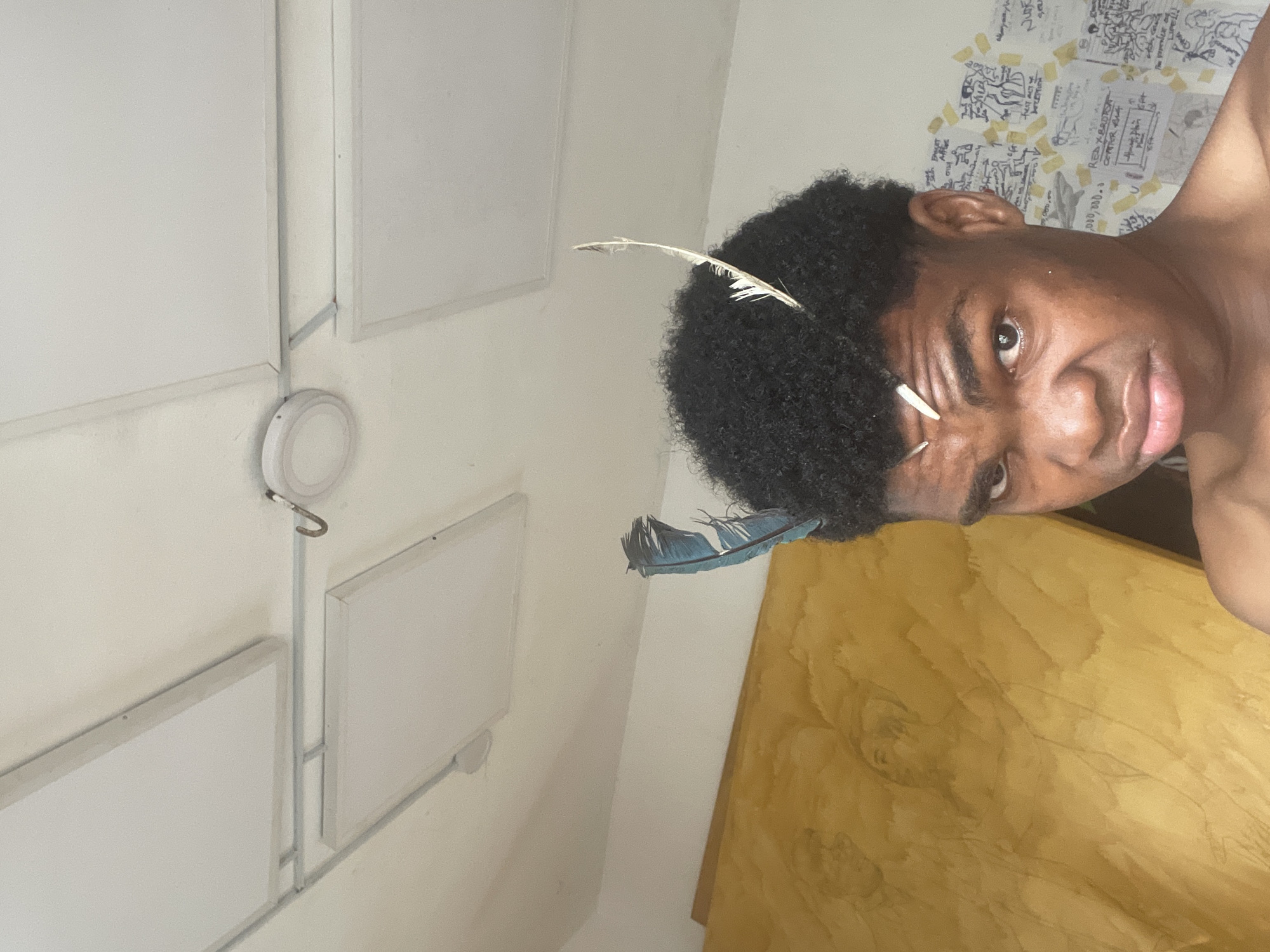 The image depicts a close-up, high-angle shot of a young Black male with dark, curly hair adorned with two feathers. One feather is blue and positioned to the left of his head, while the other is white and positioned to the right. He is shirtless, and his facial expression is a slight pout. In the background, a wall features what appear to be sketches and writings taped to it. To the left of the frame, a large, textured, yellow surface is visible, possibly a painted wall or canvas. The ceiling above has visible panels and some lighting fixtures. The scene appears to be indoors, in what could be a residential or studio space in the city of Ado, Nigeria.