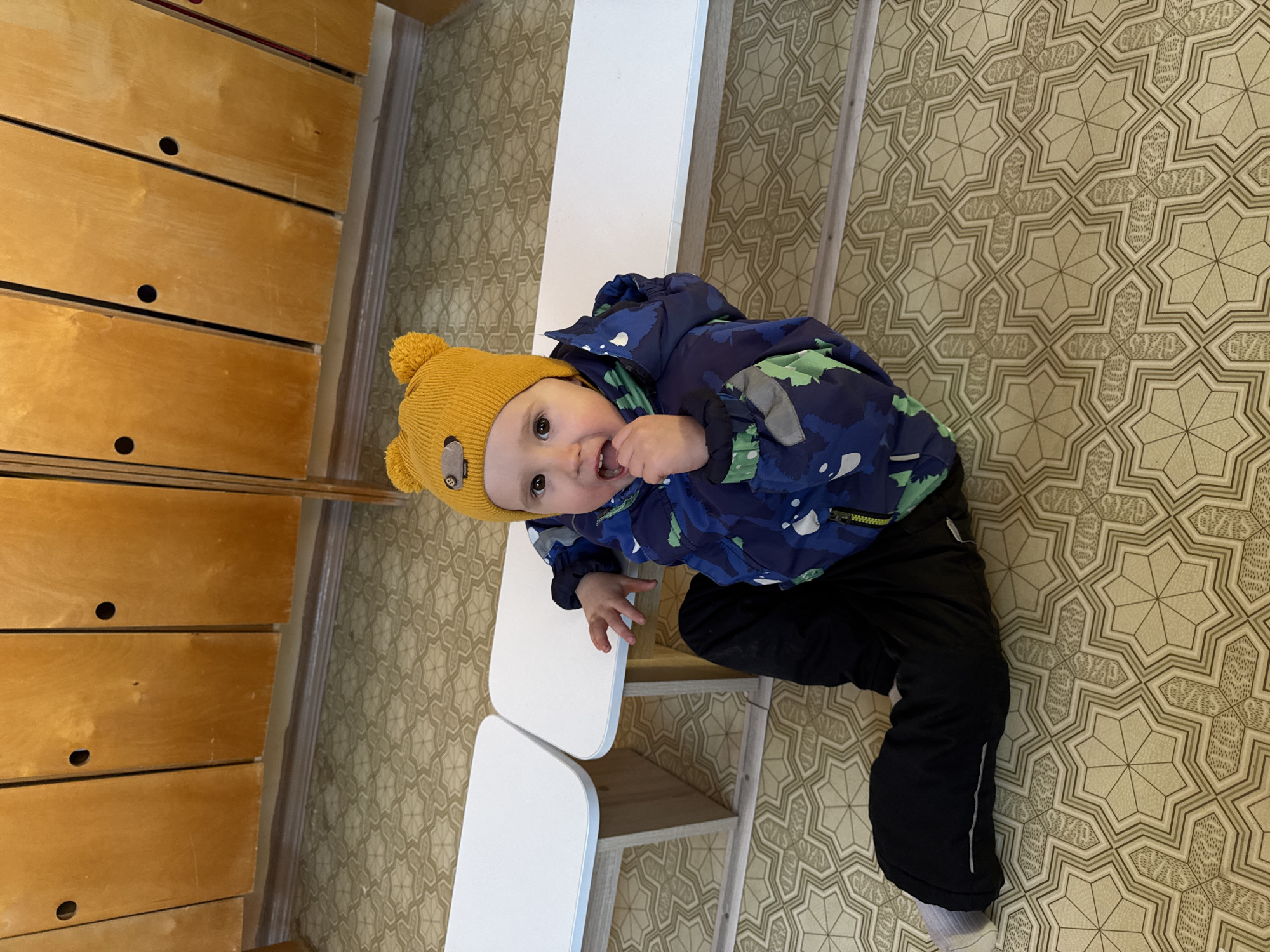 A young child wearing a yellow hat with a pom-pom, a blue and green camouflage jacket, and black pants is sitting on a white, two-tiered bench. The child is looking up at the camera and has their fingers in their mouth. The background features wooden drawers on the left and patterned linoleum flooring. There is no indication of the location being Noviy, Russia.