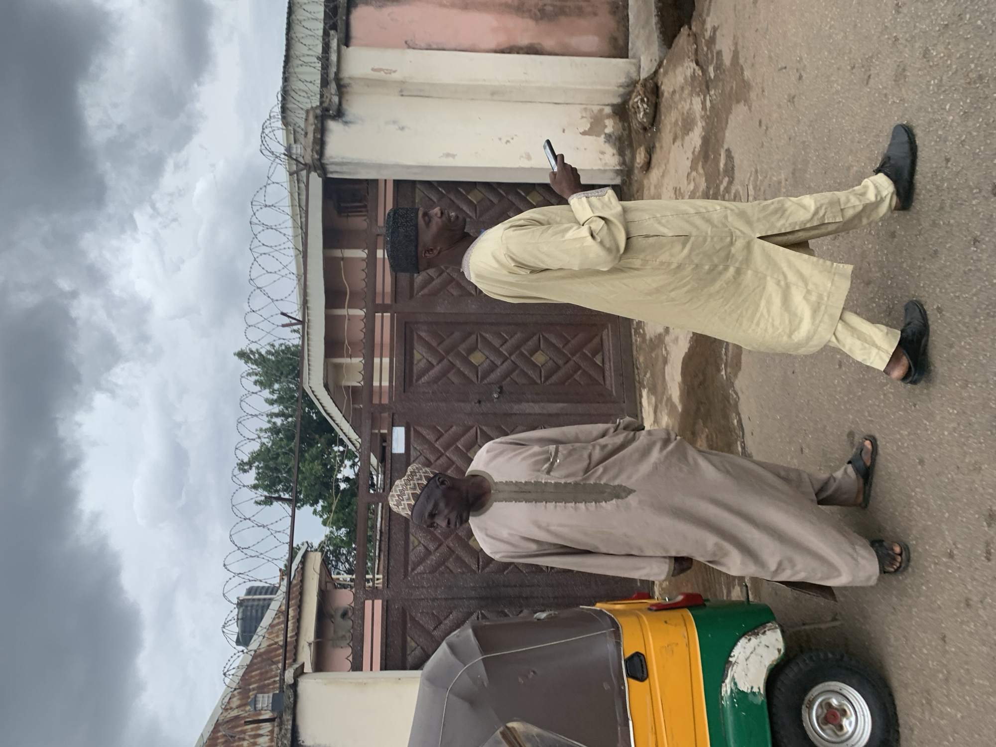 Two men are standing on a city street in Gura Top, Nigeria, next to a parked, green and yellow auto rickshaw. One man, dressed in a light beige outfit and black shoes, is looking at a cell phone he holds in his hand. The second man, in a light brown outfit and sandals, is looking down at the auto rickshaw. A building with a gate topped with barbed wire is in the background. The sky is overcast.