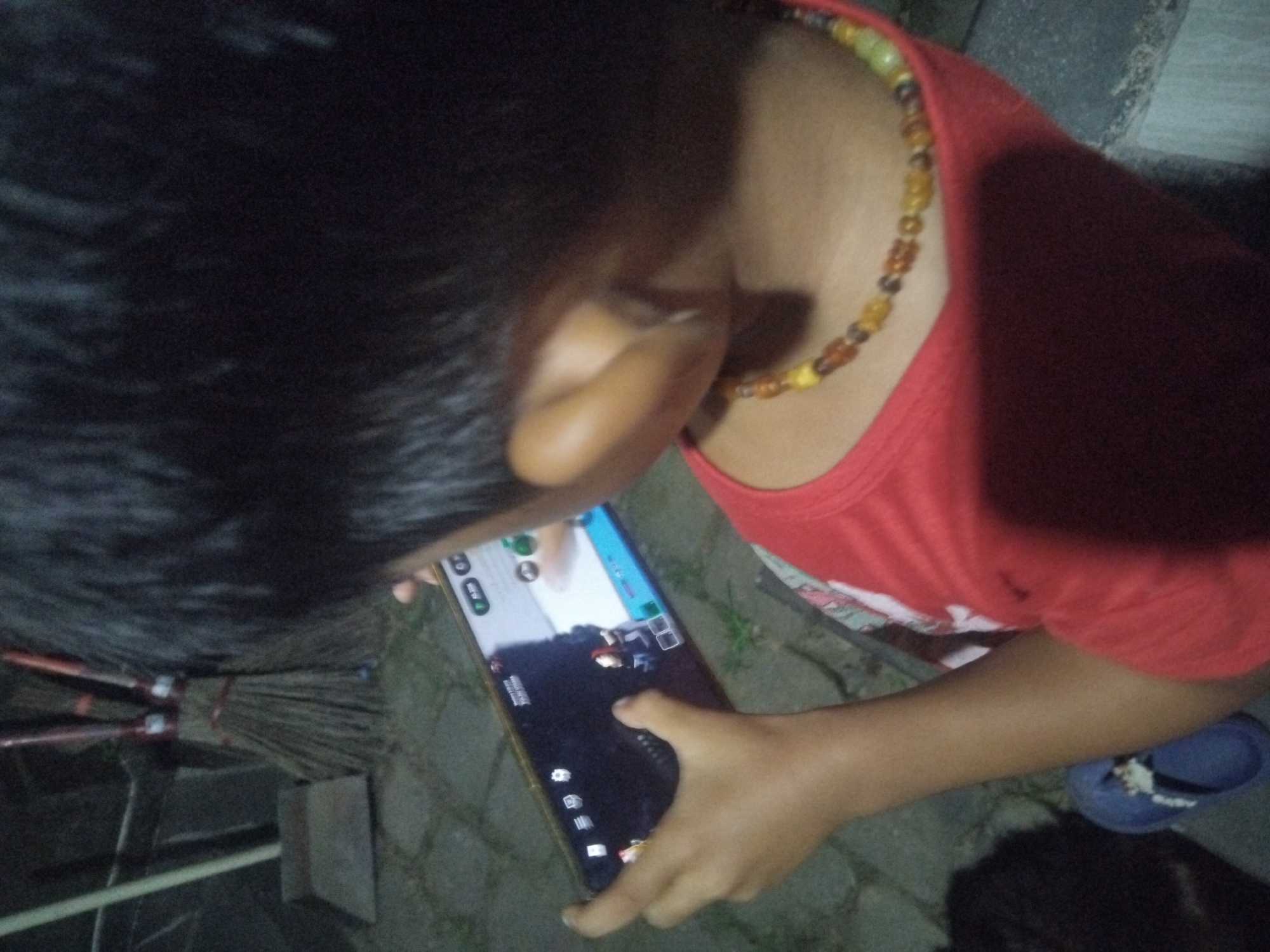 A child with dark hair and a beaded necklace is playing a video game on a smartphone. The child is wearing a red t-shirt and is partially visible from the upper chest up. The child is outdoors on a paved surface, near a broom and a sandal. The location is Unknown, Unknown.