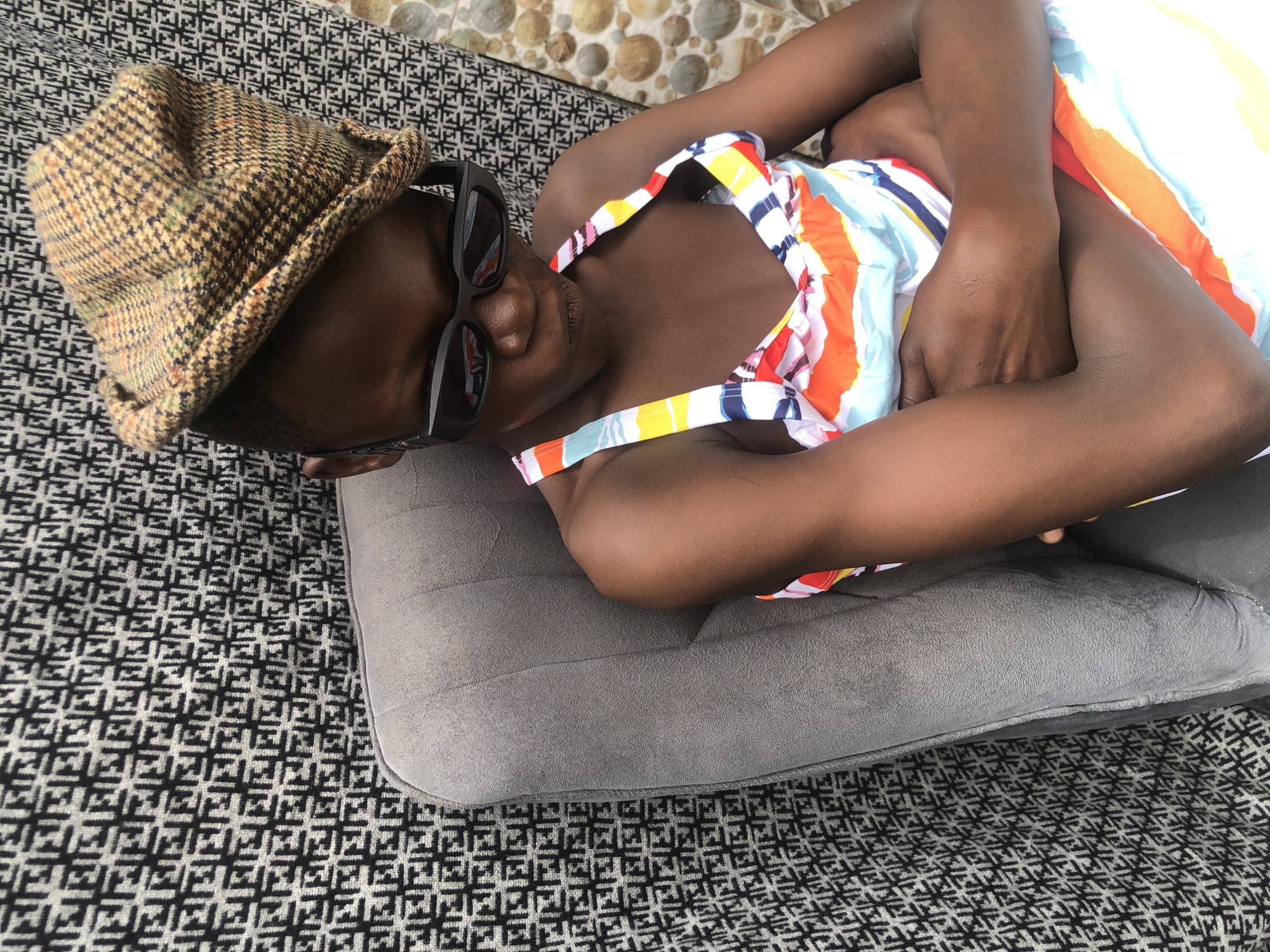 A young Black person, wearing sunglasses and a plaid flat cap, sits with arms crossed on a gray armchair. The person is dressed in a colorful, striped swimsuit. The background features a textured black and white fabric with a repeating pattern and a tiled wall. The scene appears to be outdoors, possibly in a residential area in Ado, Nigeria.