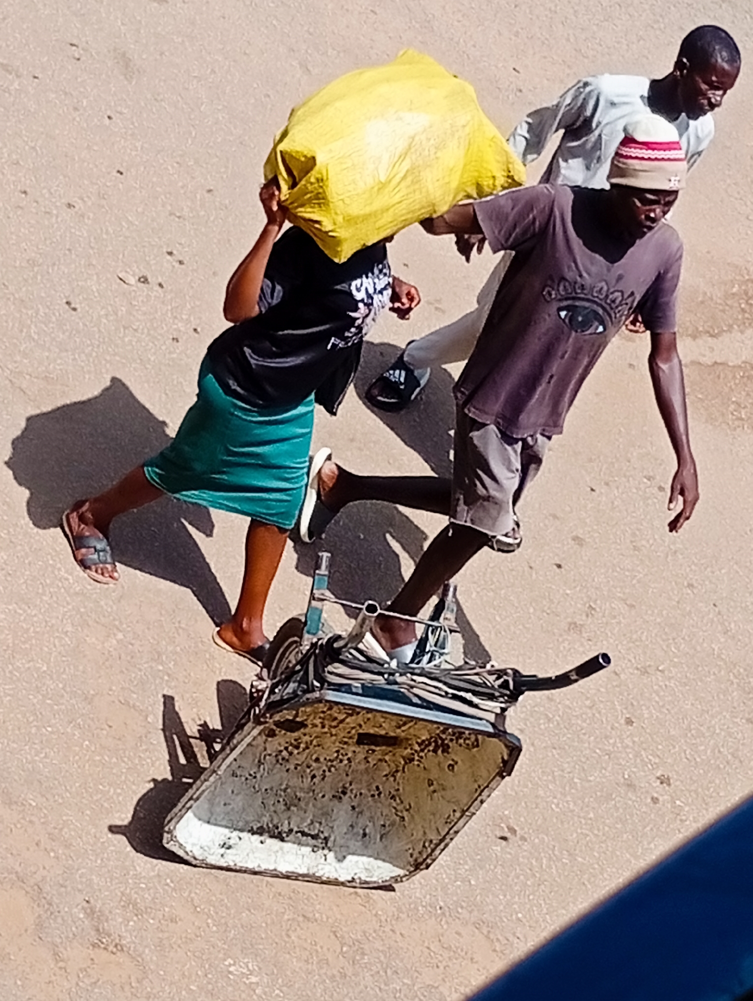 In the city of Bukuru, Nigeria, three individuals are captured in an overhead shot on a dusty, sandy surface. A person in the foreground, wearing a dark t-shirt with a stylized eye design and grey shorts, is pushing a two-wheeled cart. The cart appears to be made of metal and has a weathered, speckled texture. To their left, another individual, wearing a black t-shirt and a flowing green skirt, walks beside the cart, carrying a large, bright yellow bag over their head and shoulder. Behind them, a third person, dressed in a light-colored long-sleeved shirt and a patterned cap, walks slightly behind and to the right. The ground is a uniform sandy brown, suggesting an outdoor environment. The lighting indicates it is daytime, with distinct shadows cast by the individuals and the cart. The overall impression is one of labor or transport, with the three individuals working together.