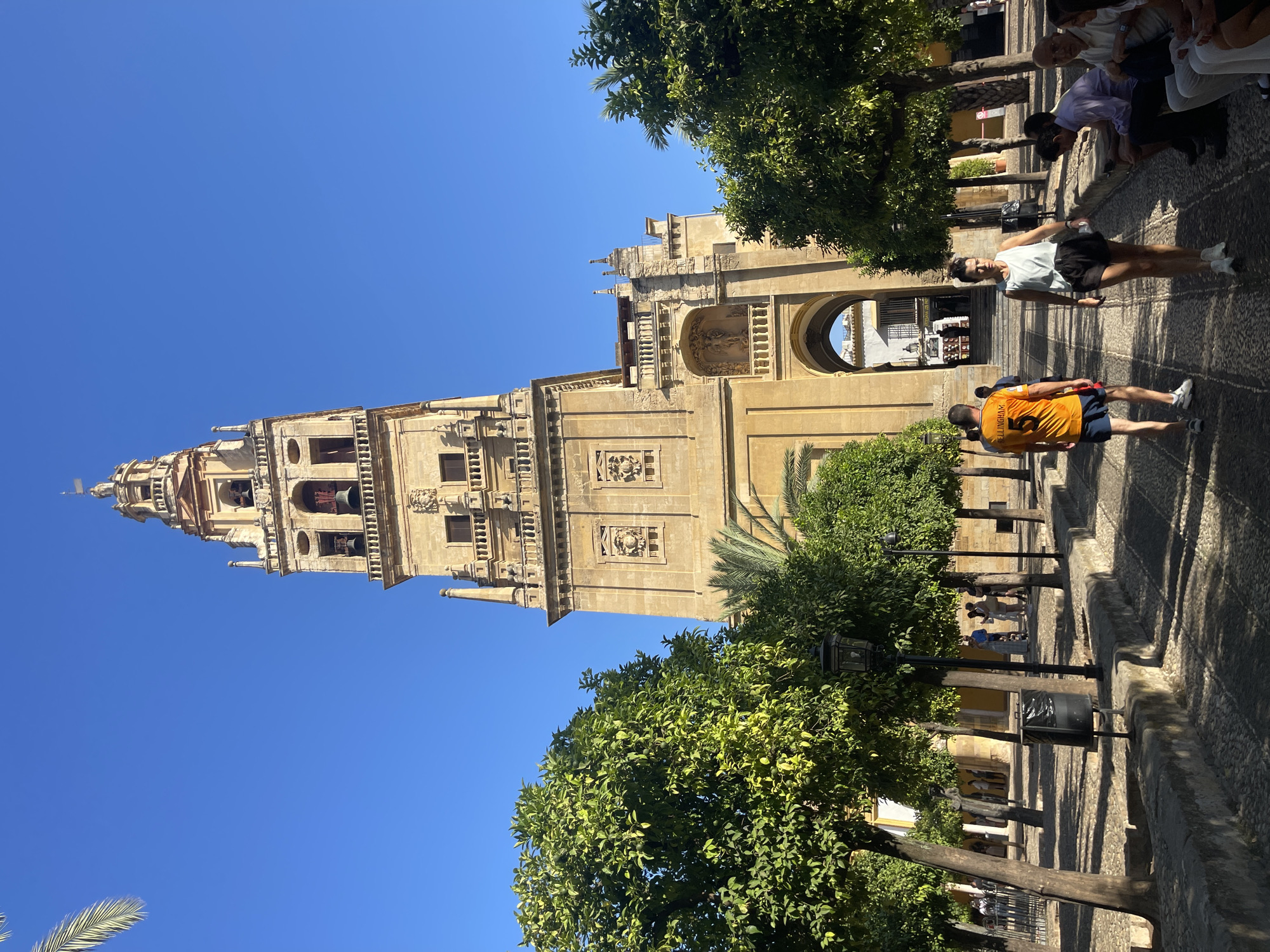 The image shows the Mosque-Cathedral of Córdoba, Spain, specifically its bell tower (La Giralda), and the surrounding courtyard. The bell tower, a tall, square structure made of beige stone, dominates the background. It features multiple levels with arched windows and decorative carvings. In the foreground, a paved courtyard with cobblestones is lined with orange trees, casting dappled shadows. Several people are present in the courtyard. Two individuals are walking towards the viewer, one wearing an orange shirt with the number 5 and the other a white tank top and black shorts. Other individuals are seated on a low stone wall on the right side of the image, some looking at their phones. In the middle ground, people are seen walking through an arched entryway. The sky is clear and blue.