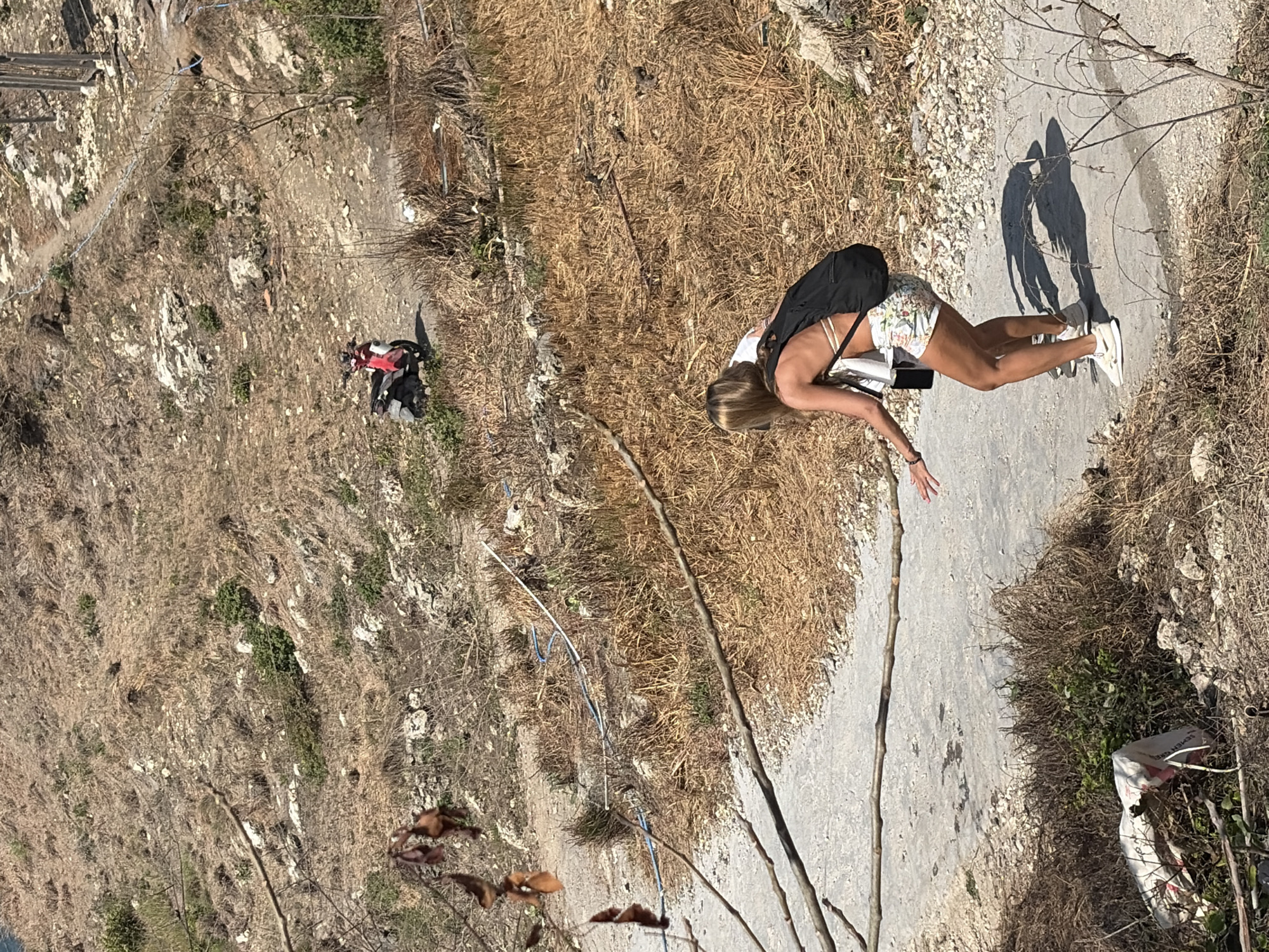 A woman wearing a white bikini top, floral shorts, and sneakers, with a black backpack, bends down on a gravel path in a dry, hilly area. In the background, a red and white scooter is parked on a path further up the hill, and two brown goats are visible on the left side of the frame. The environment is characterized by dry grass, sparse vegetation, rocks, and a rough, unpaved path. The scene appears to be in a natural, undeveloped setting, likely within the landscape of Nusa Penida, Indonesia, known for its rugged terrain and coastal views.