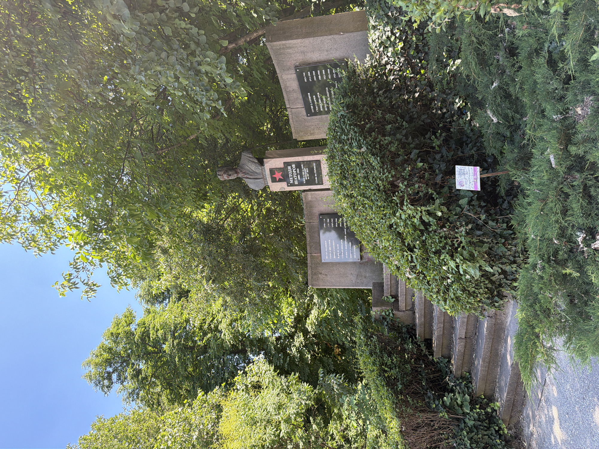 The image depicts a monument in a park-like setting. The monument features a bronze bust of a man atop a pedestal, which is adorned with a red star. To the right of the bust, a curved concrete structure holds several plaques with names and dates. To the left of the pedestal, another concrete structure supports a plaque. Stone steps lead up to the monument, flanked by lush green bushes and trees. The ground is covered with gravel. A small informational sign is visible in the foreground, partially obscured by greenery. The sky is clear and blue. This is the monument to partisans in Pazardzhik, Bulgaria, commemorating individuals who fought against fascism during World War II.