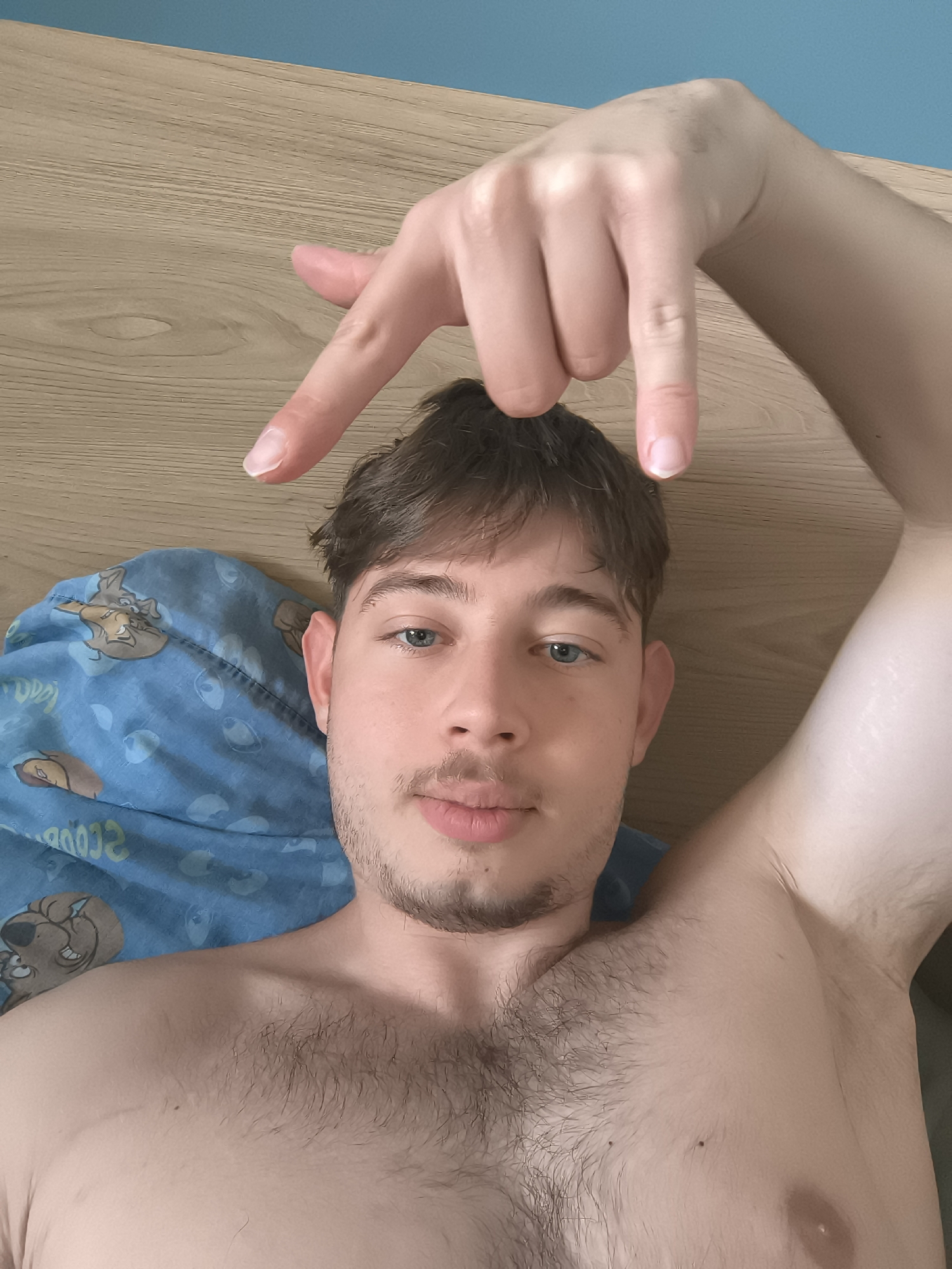 A shirtless young man with blue eyes and brown hair is lying on his back, possibly in a bed, indicated by the blue patterned blanket and wooden headboard visible. He is holding his right hand above his head, with his index and middle fingers pointing downward towards his head. The location of the scene is not discernible from the image.