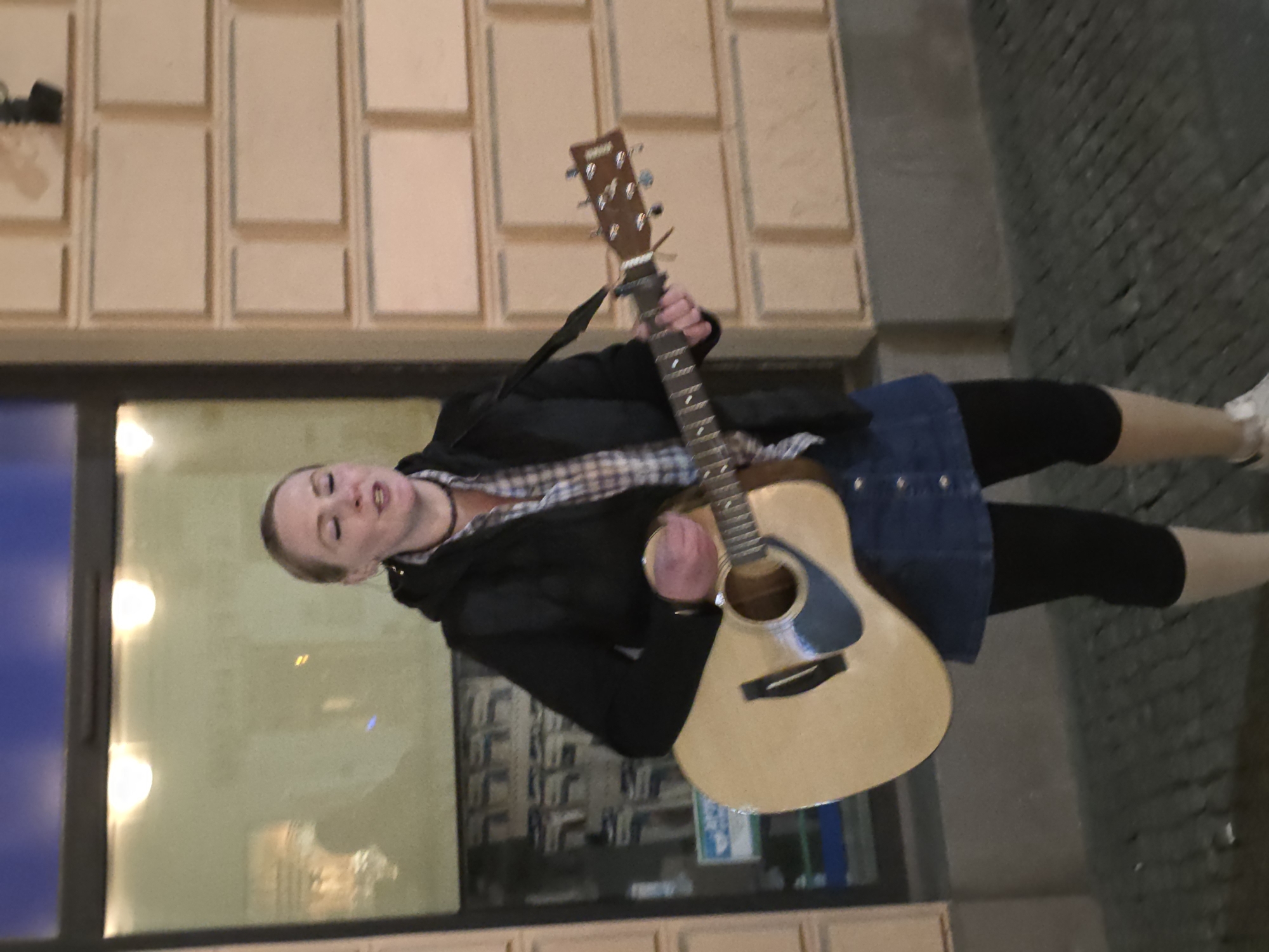 A woman is standing on a cobblestone street in Warsaw, Poland, holding a guitar. She is wearing a black jacket, plaid shirt, denim skirt, black leggings, and beige socks. The woman is positioned in front of a building with a display window featuring illuminated lights. The building facade features a brick pattern.