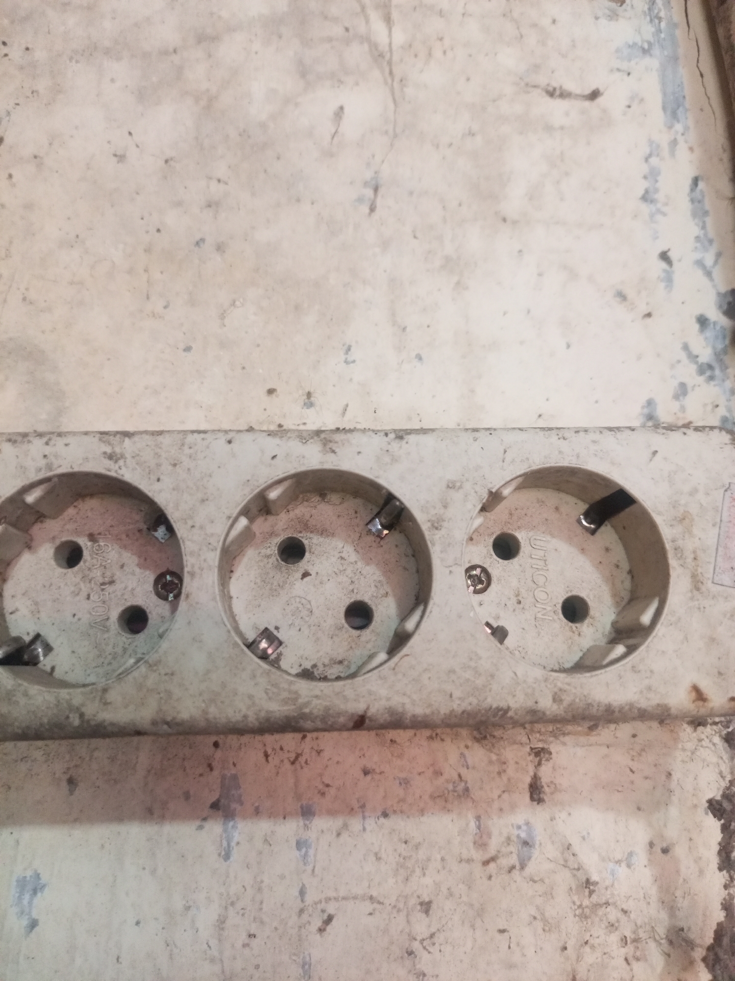 The image shows a close-up of a dirty, multi-outlet power strip with three visible sockets. The power strip is mounted on a worn, off-white surface that appears to be a wall or a similar structure. The sockets are dusty and have visible metal contacts. One socket has the text "UTICON" imprinted on it. The overall environment appears to be indoor and somewhat neglected, with dust and grime covering the surfaces. The location is specified as Lhokseumawe, Indonesia.
