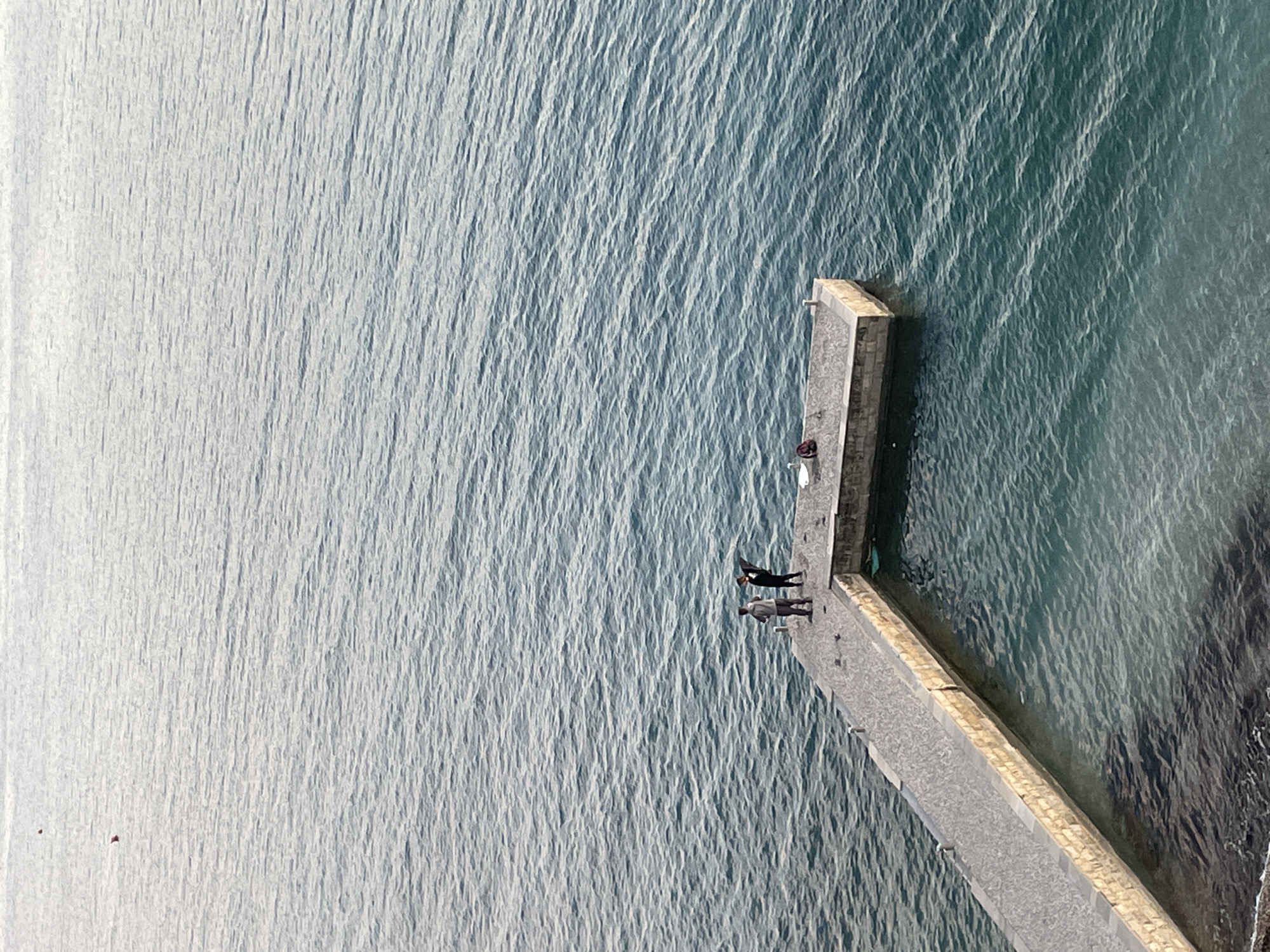The image displays an elevated view of an L-shaped stone pier extending into calm, blue-green water, identified as located in Budva, Montenegro. The pier features a longer segment paralleling the shore and a shorter, perpendicular segment extending into deeper water. Two individuals are positioned on the far end of the perpendicular pier segment. The person on the left wears a grey top and dark trousers, oriented towards the right. The person on the right wears dark attire, with arms or a garment extended laterally, facing away from the viewer. A red and black bag and a white bucket are situated on the pier surface near the second individual. Two small red buoys are visible on the water's surface in the upper-left background. The water exhibits gentle ripples and variations in color consistent with depth.