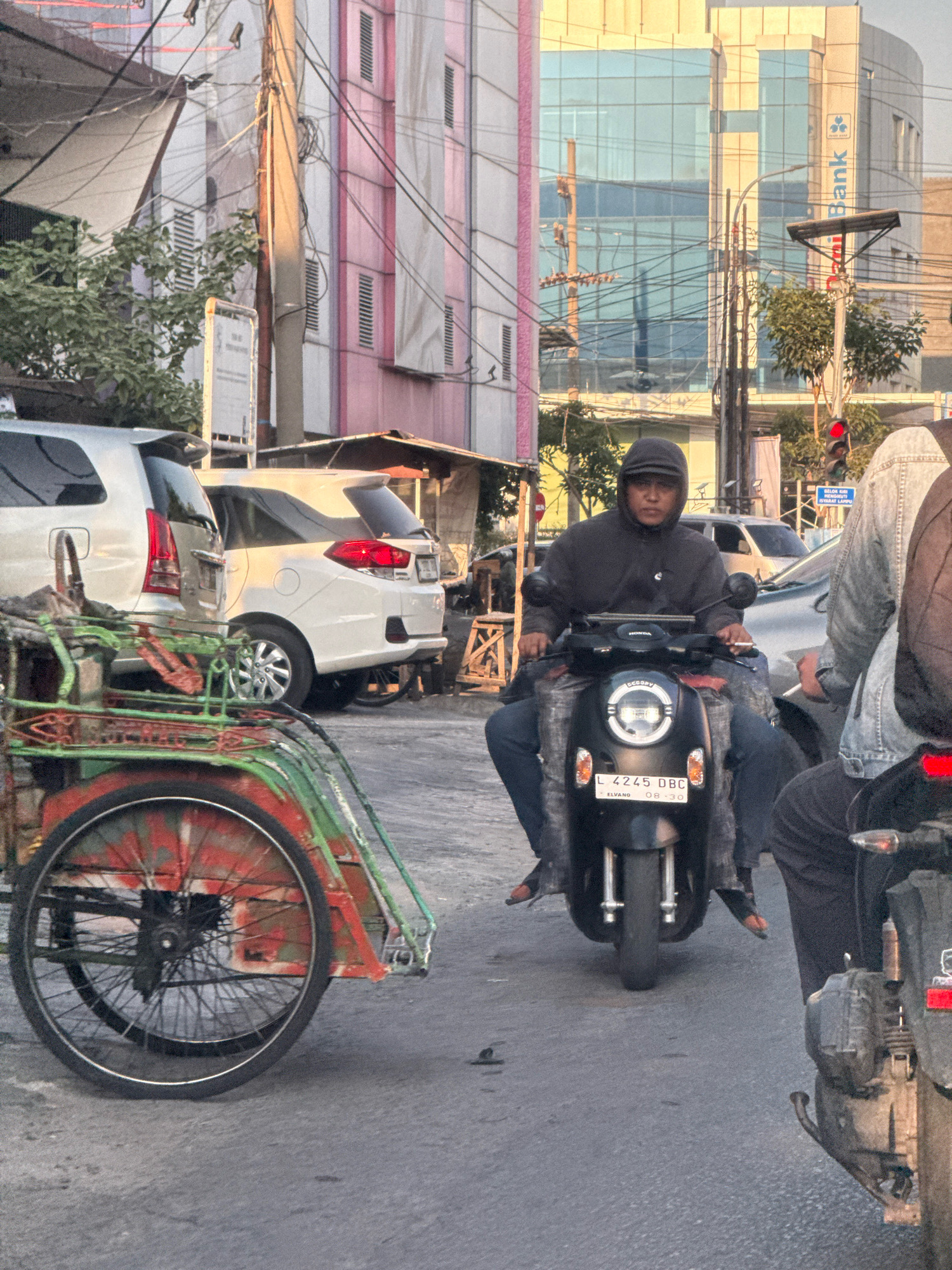 In Surabaya, Indonesia, on a city street, a man wearing a black hooded jacket and a cap rides a black Honda scooter. He is looking forward, and his face is partially obscured by his hood. He has a cigarette in his mouth. The scooter has a license plate that reads "L 4245 DBC". He is wearing blue jeans, and his feet are visible, wearing shoes. The scooter has a white headlight and a digital display. Behind the scooter, another person is partially visible, wearing a denim jacket. In the foreground, to the left, a pedicab with green and red metal frame and a large black wheel is parked. In the background, there are buildings and trees. One building is modern with a glass facade and the logo of Bank Mandiri is visible. Another building has a pink and white facade. There are power lines crisscrossing the scene. The lighting suggests it is late afternoon, with the sun casting long shadows.