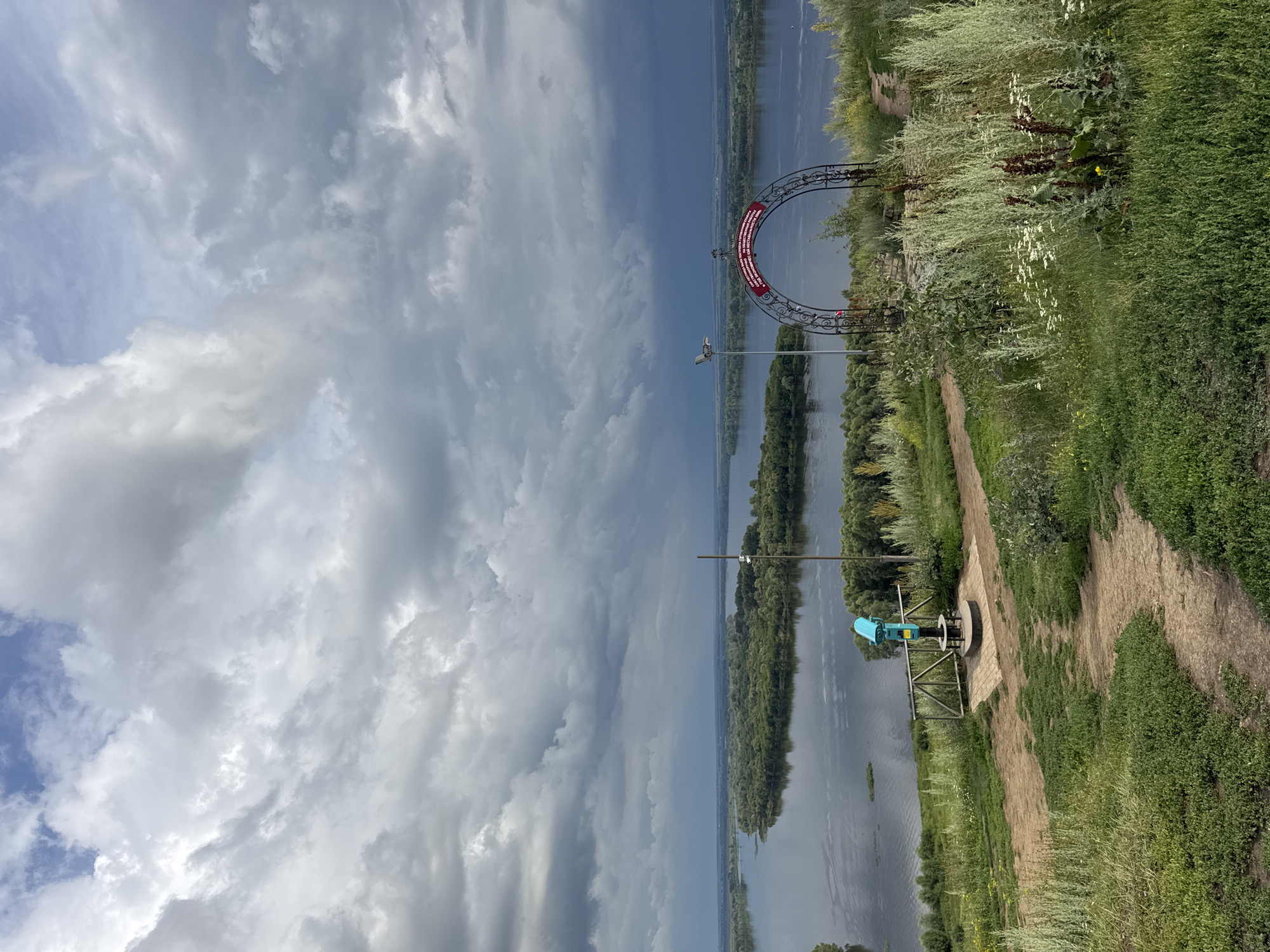 This is a wide shot of the Volga River in Yelabuga, Russia. The sky is dramatic with dark, stormy clouds covering most of the upper portion of the image, with a small patch of blue sky visible on the top left. The river is wide and calm, with a lush green island in the middle of it. On the bank, in the foreground, is a grassy hill with a dirt path winding through it. To the left, there is a blue viewing telescope on a pedestal, behind a metal fence. To the right, there is an ornate metal archway with red lettering on it. The archway is decorated with what appears to be a weather vane on top. The overall scene is one of natural beauty with hints of human presence.