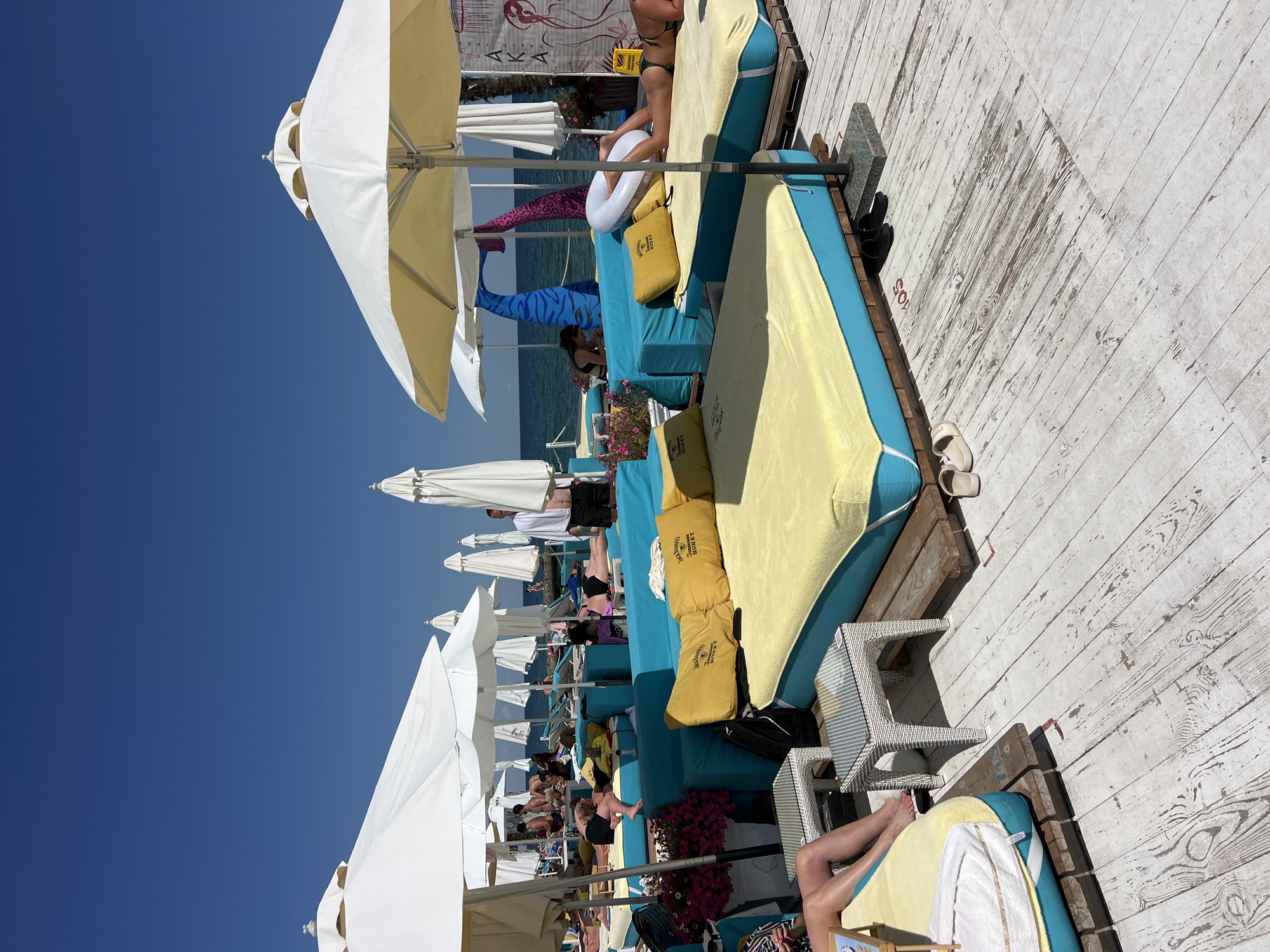 This is a wide shot of a beach club on a sunny day in Odesa, Ukraine, with a clear blue sky and the sea in the background. The foreground features a wooden deck with several large, low-lying daybeds. These daybeds are upholstered in bright blue with thick yellow cushions and pillows, some of which have "Jack Daniel's Honey" branding. White beach umbrellas are erected over some of the daybeds.
In the mid-ground, several people are relaxing on the daybeds, some sunbathing and others chatting. A man in a white shirt and black shorts stands on the right side of the frame, appearing to be an attendant. Beyond the daybeds, more beach umbrellas and seating areas are visible, as well as the blue sea. The overall impression is a scene of leisure and relaxation at a resort or beach club.