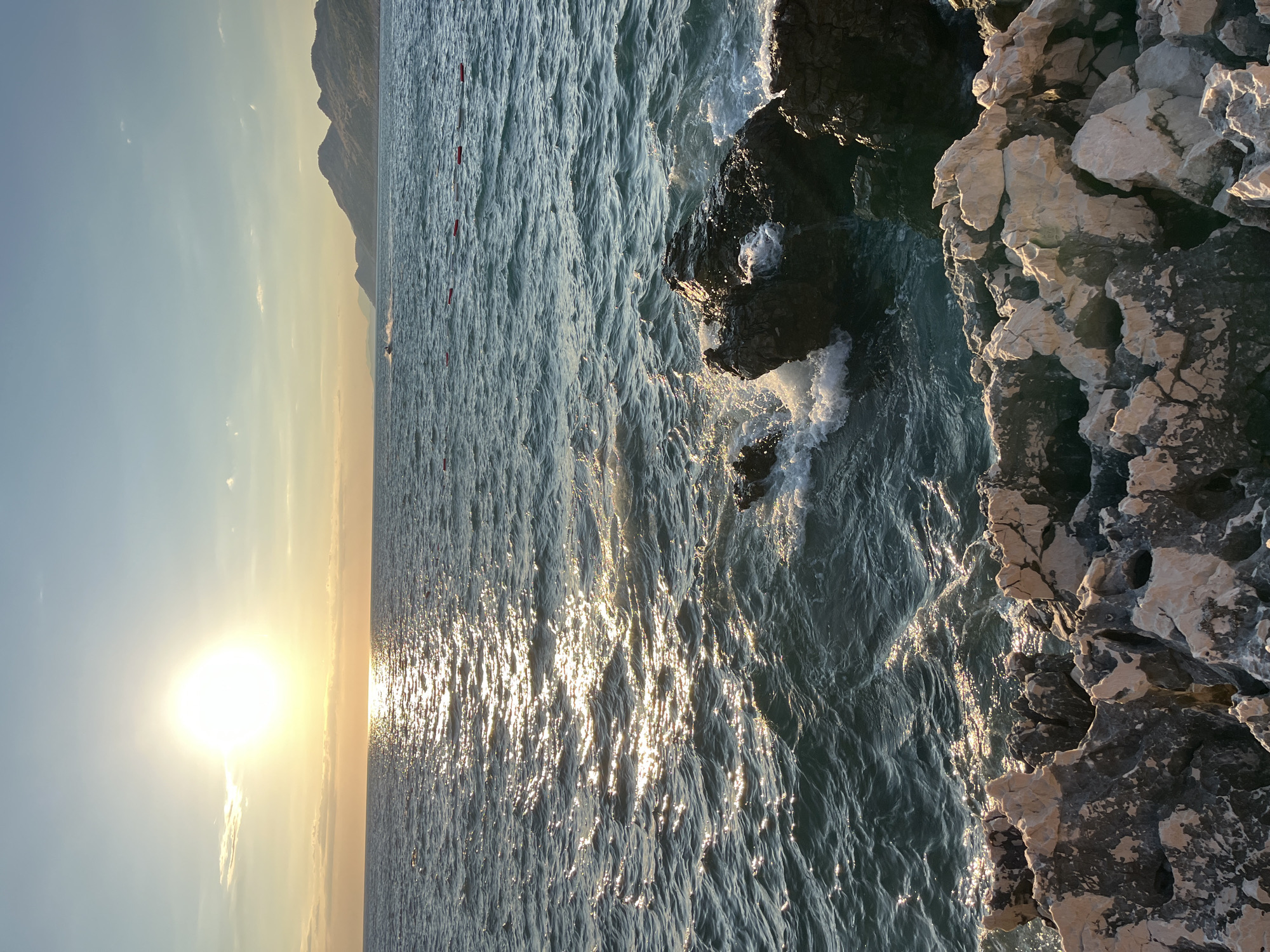 The image depicts a coastal scene at sunset, likely in Pečurice, Montenegro. The sun is low on the horizon, casting a warm glow and creating a shimmering reflection on the rippling, dark blue sea. In the foreground, rugged, light-colored rocks are visible, some of which are wet from the waves crashing against them. Further out, the water is dotted with a line of red buoys. In the distance, a hazy line of mountains forms the backdrop, and a jet ski can be seen creating a spray as it moves across the water. The sky transitions from a pale yellow near the horizon to a clear blue higher up, with a few wispy clouds.