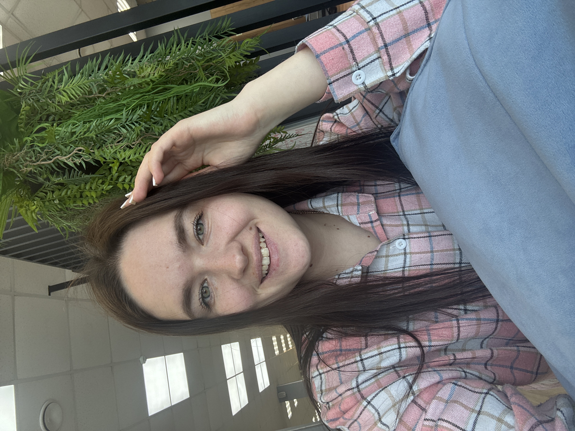 A young adult female, with long brown hair, green eyes, and visible freckles, is depicted smiling directly at the camera. She wears a pink, white, and blue plaid button-up shirt. Her right hand, adorned with white-tipped nails, is raised to the right side of her head, touching her hair. A light blue fabric object, possibly a blanket or cushion, is visible in the lower right foreground. The background features a drop ceiling with square fluorescent light panels and a circular white fixture (likely a smoke detector or sprinkler head). To the right, a black slatted structure and artificial green foliage are present, suggesting an indoor commercial or public setting, possibly in the city of Chaikovsky, Russia.