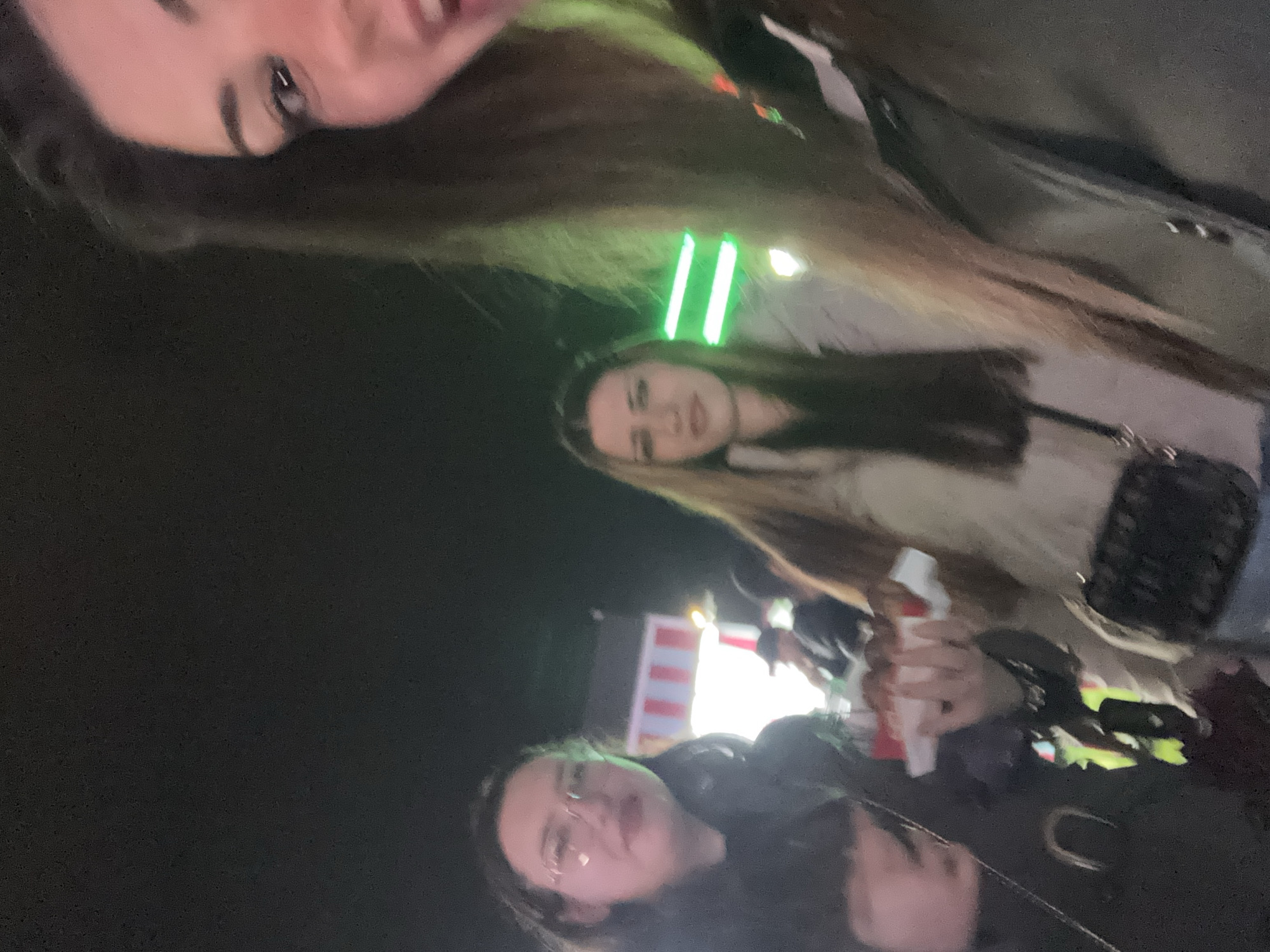 Three women are in the frame, likely taking a selfie at night in what appears to be an outdoor event. The woman at the top of the frame is partially visible, showing her face and long brown hair. The woman in the middle has light-colored hair and is wearing a light-colored jacket with a dark purse strap across her body. The woman at the bottom of the frame is wearing glasses and dark clothing. In the background, there are bright lights and a building with vertical red and white stripes, possibly a food stand or vendor booth. It is difficult to determine the location in Sofia, Bulgaria without more specific identifiers.
