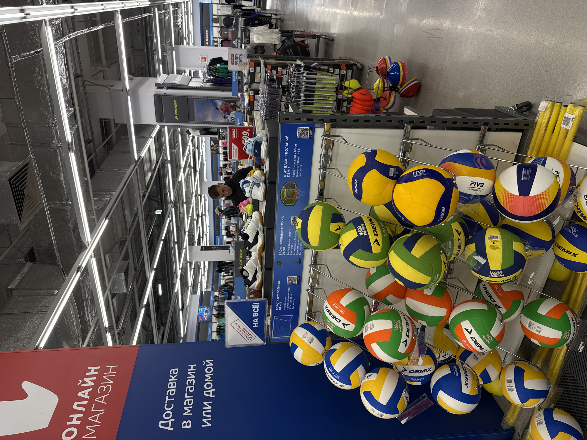 The image depicts an indoor scene, seemingly within a retail store in Krasnoyarsk, Russia. The store features high ceilings with exposed ductwork and fluorescent lighting. A variety of sports equipment is on display, including volleyballs of varying colors and designs, basketball equipment, and soccer goals. A person is seen near a display of athletic shoes, looking at the merchandise. A blue and red sign with text in Russian advertises online shopping and home delivery. The products are labeled with the "Demix" brand name. Some of the volleyballs are branded with "FIVB APPROVED" logos. There are also red/orange safety cones visible in the upper right section of the image. The scene appears to be a sports section of the store, focusing on team sports equipment.