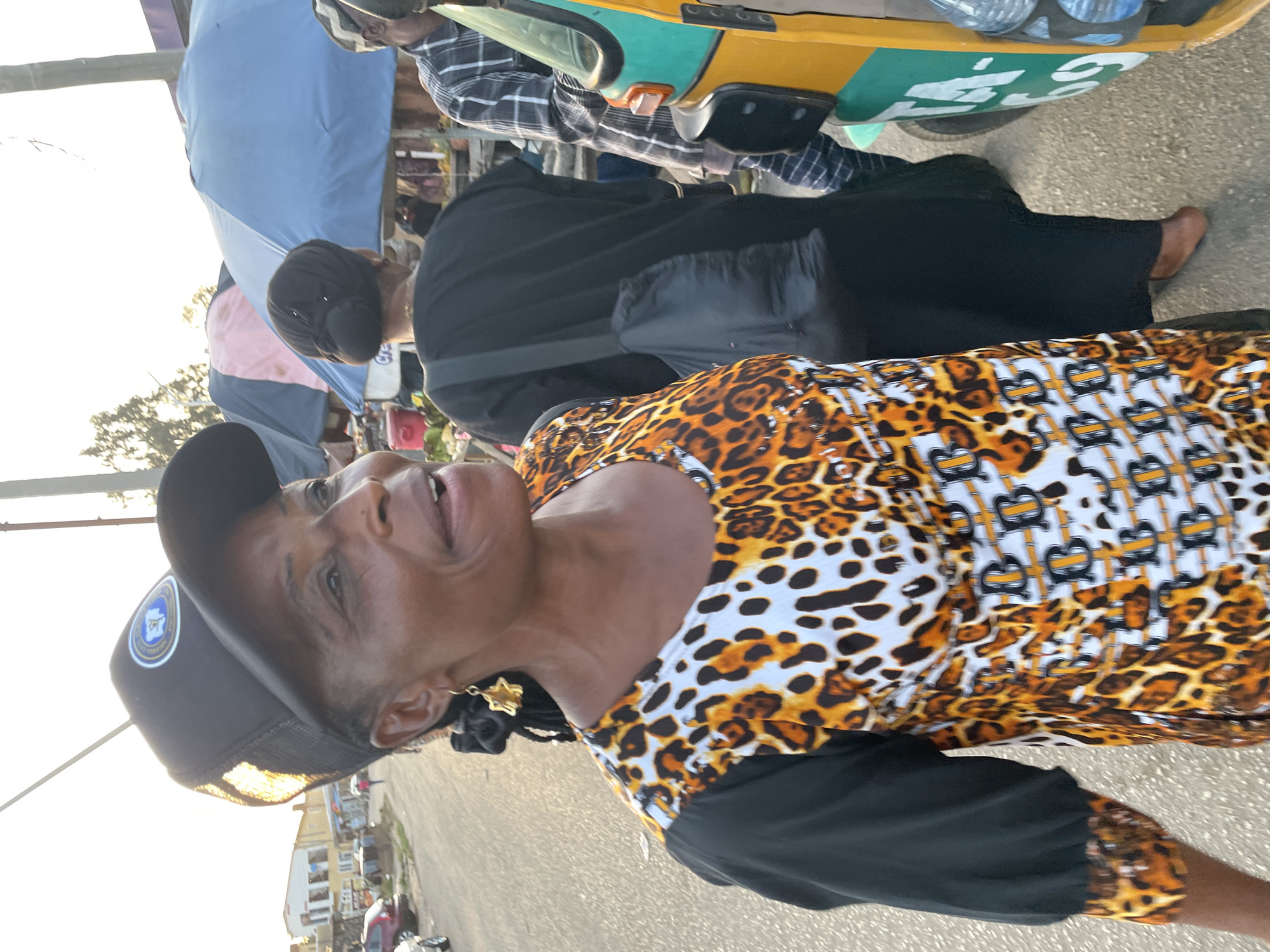A middle-aged woman stands on a street in Jos, Nigeria. She wears a black baseball cap, a patterned animal print shirt with black sleeves, and gold star earrings. Her hair is styled in braids. Behind her are other people, some under a blue awning. A yellow vehicle with green accents and water bottles strapped to the back is present. The street is paved. Buildings are visible in the background, and the sky is light.