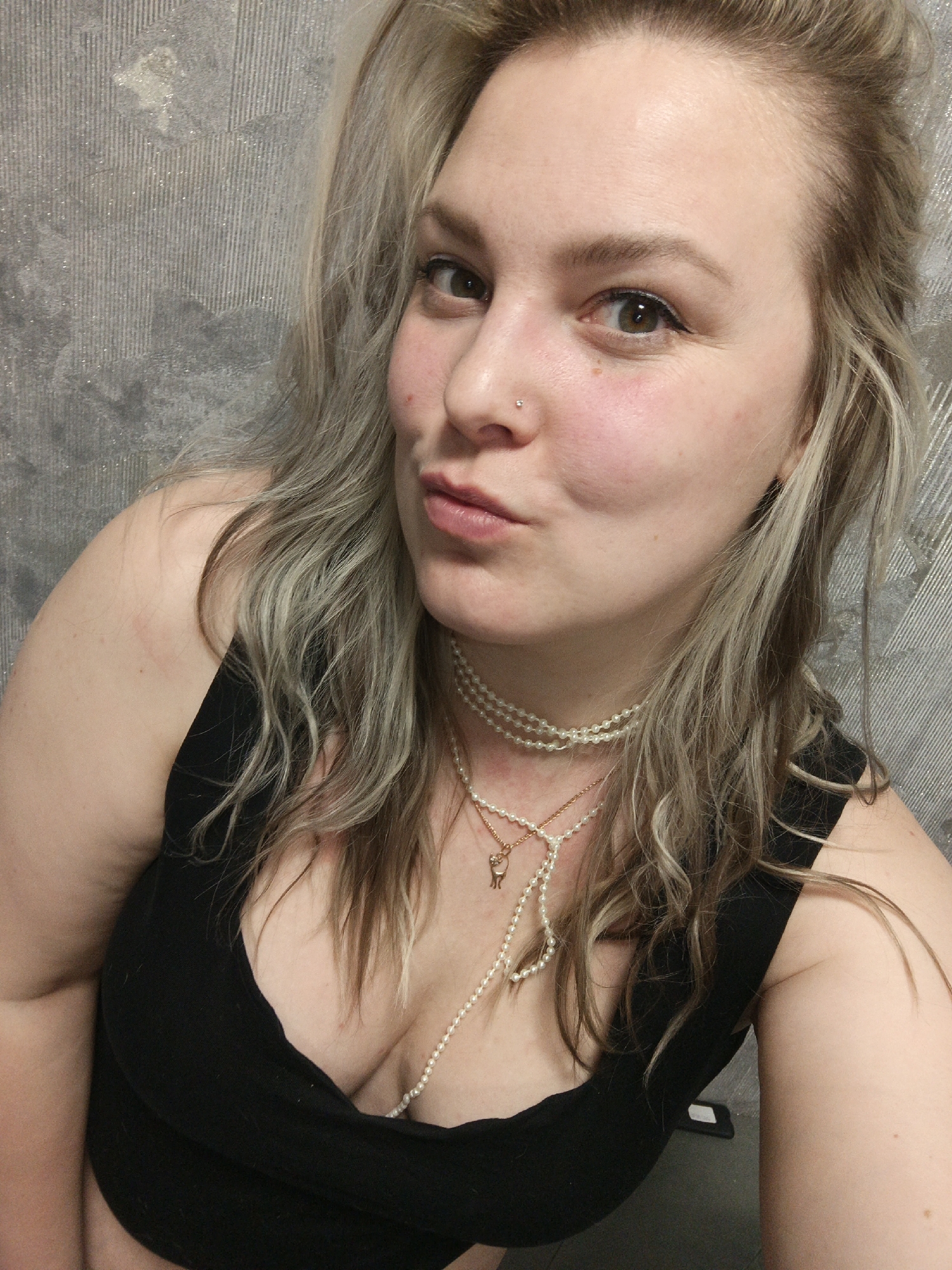 A blonde-haired woman with fair skin is posing for a selfie. She is wearing a black low-cut top and multiple pearl necklaces, one with a gold pendant. Her lips are pursed in a kiss. A nose piercing is visible on her left nostril. The background appears to be a gray textured wall. It is impossible to confirm any location or city details based on the visual information.