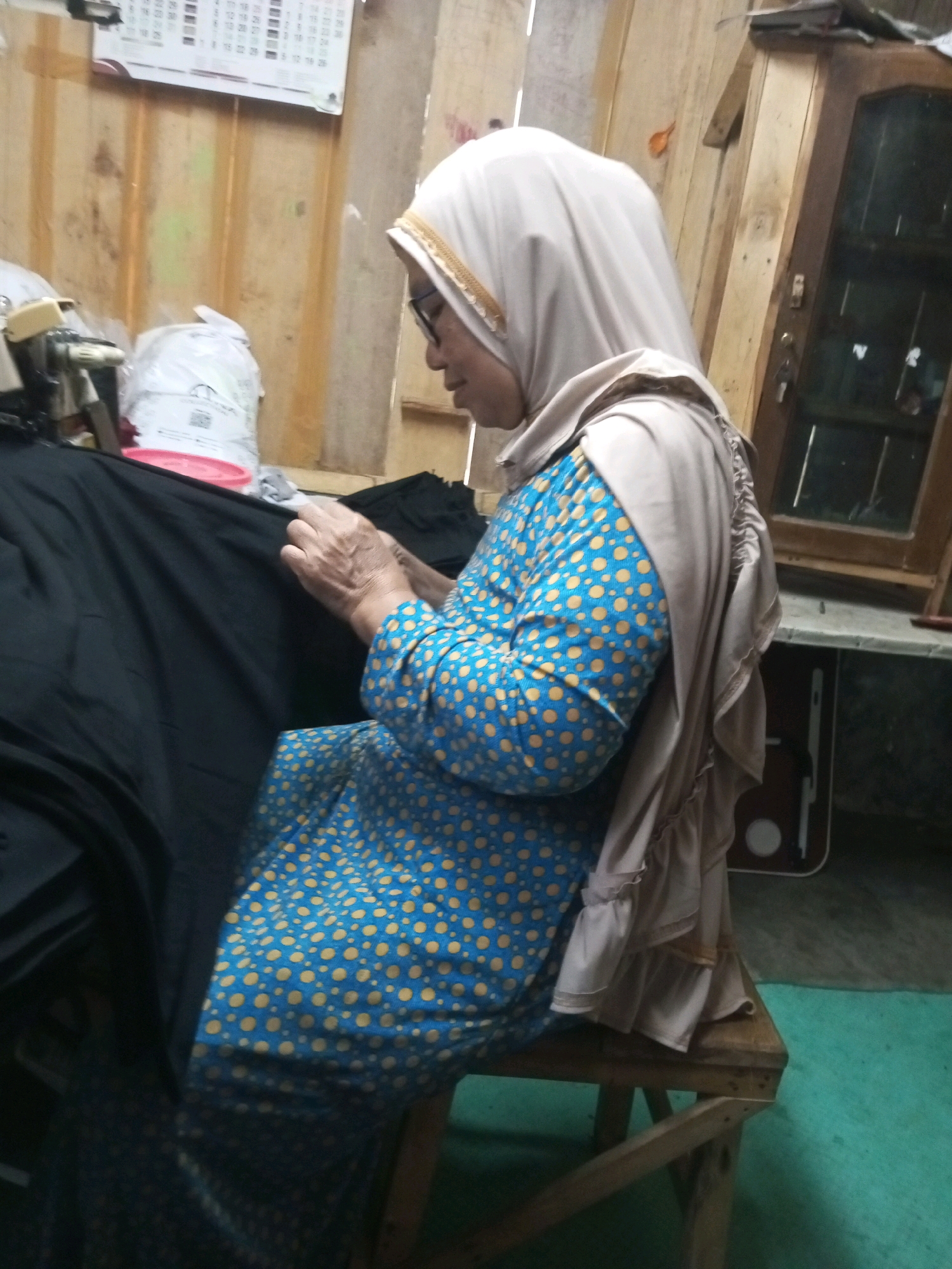 In Kalibeber, Indonesia, a woman wearing a light brown hijab and glasses is sitting on a wooden stool. She is dressed in a blue and yellow polka dot dress and a light brown ruffled scarf. Her hands are engaged in what appears to be sewing on a large piece of black fabric spread across her lap. To her left, there are several items including a white object that might be a sewing machine, a white plastic bag, and a pink circular object. The background consists of wooden walls and a wooden cabinet with a glass door. A calendar hangs on the wall behind the woman. The floor is covered with a greenish material.