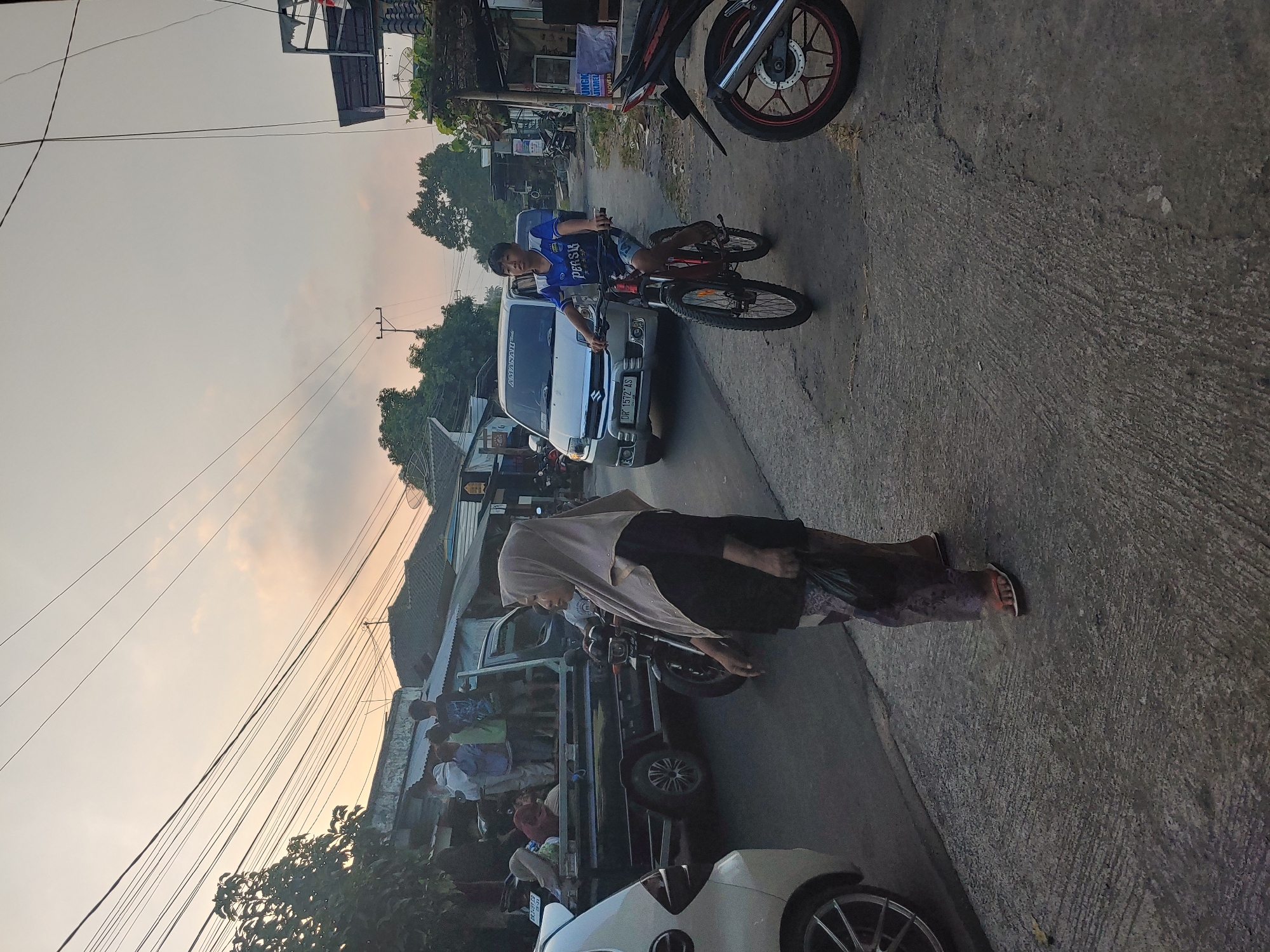 In Kembang Kerang, Indonesia, a street scene unfolds at dusk with a muted sky. A young boy rides a red bicycle down a paved road, wearing a blue t-shirt with white lettering and dark shorts. To his left, a silver van with an Indonesian license plate (DR 1572 AS) is stopped. On the right side of the road, a motorcycle is parked. In the foreground, a woman wearing a light brown hijab and a patterned skirt walks towards the left, carrying a dark plastic bag. Behind her, a black pickup truck is loaded with several people, some standing, and a woman wearing a pink headscarf is seated. Various buildings and trees line the street, with numerous overhead wires crisscrossing the sky.