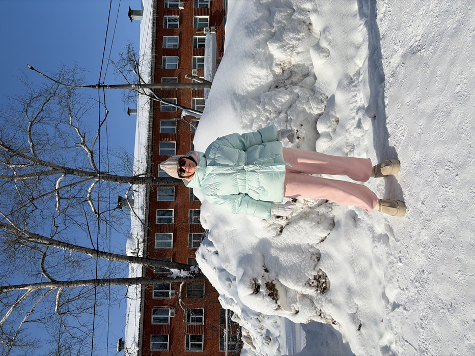 This image depicts an urban street scene in Chaikovsky, Russia, during winter. A single female individual stands centrally, facing the camera on a snow-covered surface. She is dressed in a light pink knitted beanie, dark sunglasses, a light mint green belted puffer jacket with a visible pink hood lining, light pink wide-leg trousers, and light tan platform boots with intricate gold-colored trim around the ankles. White gloves cover her hands. Large, irregular mounds of compacted snow, showing embedded dirt and ice, flank the subject. In the background, a multi-story building constructed of red brick features numerous rectangular windows with white frames. Its roof is partially covered in snow, and multiple prominent icicles hang from its eaves. Several bare deciduous trees with dark trunks and branches are visible both in front of and behind the building. A dark utility pole with a street light fixture is positioned on the right side of the frame. The sky is clear and bright blue, indicating a sunny day.