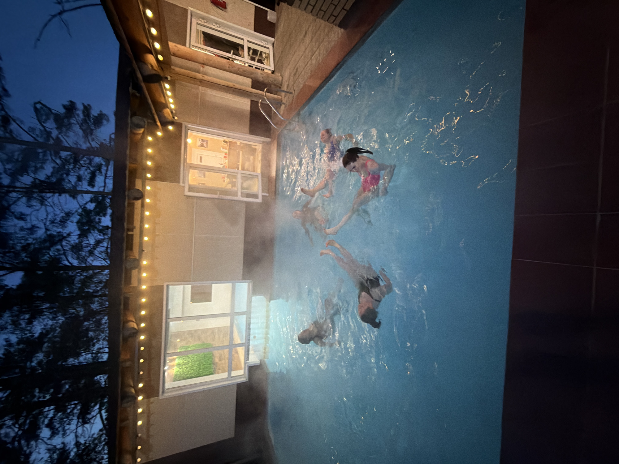 The image captures an evening or nighttime scene at an outdoor swimming pool, located beside a building. Several individuals, primarily children or young adults, are depicted enjoying themselves in the water. Visible steam rises from the pool's surface, indicating that the water is heated, likely a hot tub or a pool in cooler temperatures.
The building adjacent to the pool, possibly a house or guesthouse, features multiple windows that emit warm light. Along the eaves or a structural beam of this building, strings of glowing lights are strung, adding a festive and inviting ambiance to the scene. A pool ladder is visible on the edge of the pool.
Beyond the illuminated area, the dark sky of dusk or night is visible, along with the silhouettes of numerous tree branches and foliage, suggesting a natural, possibly wooded, setting. The pool water reflects the surrounding lights, creating shimmering patterns. The individuals in the pool appear to be swimming, floating, and generally enjoying the activity. No visible text is present in the image.