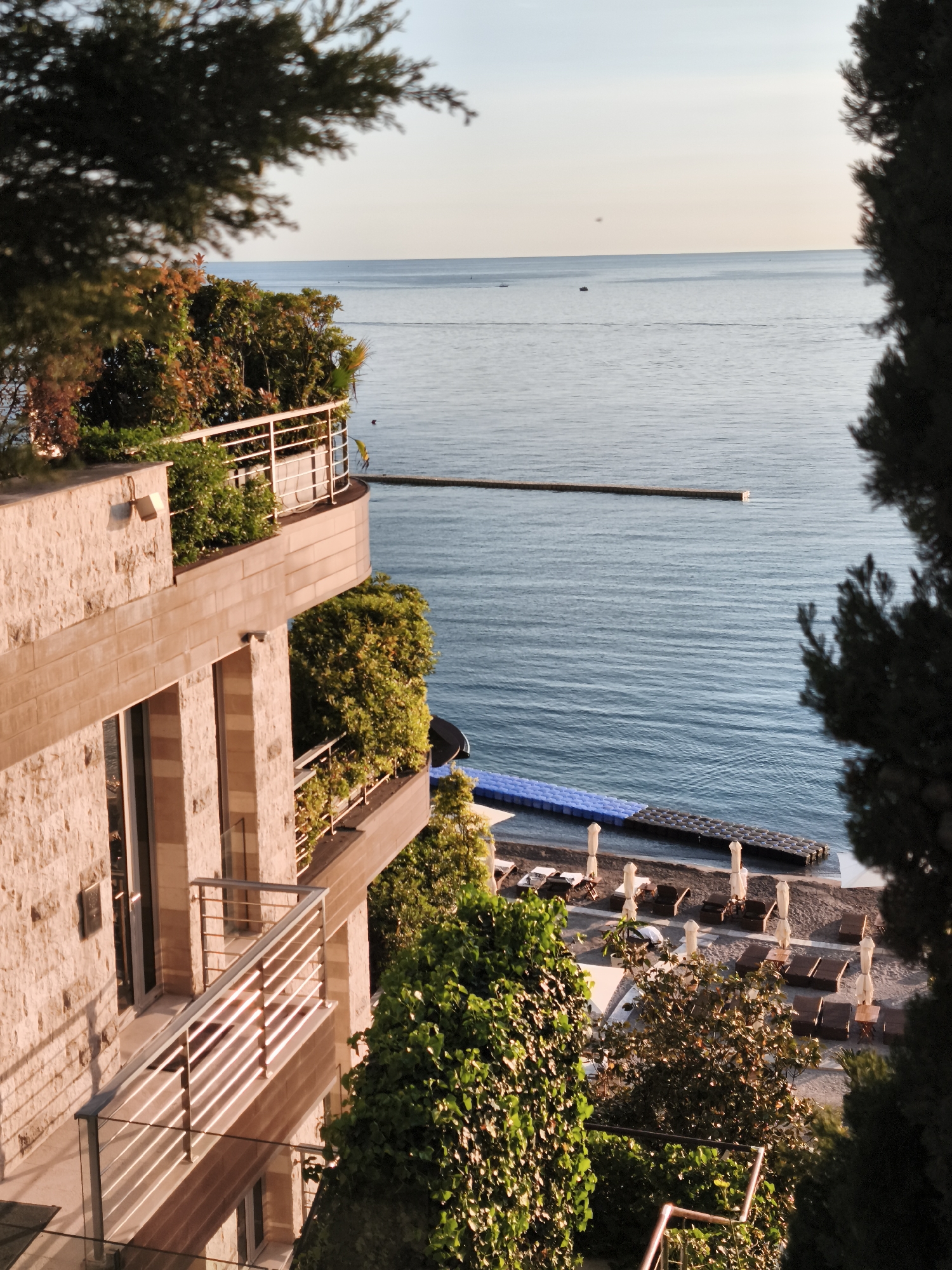 This picturesque, elevated view captures a serene coastal scene, likely from a resort in Budva, Montenegro, during the golden hour. The soft, warm light of late afternoon or early morning bathes the environment, creating a tranquil atmosphere.
In the foreground and to the left, a modern multi-story building with a light-colored, textured stone facade dominates. Its balconies, adorned with lush green plants, feature sleek metal and glass railings that reflect the soft sunlight. A light fixture is visible on one of the upper walls.
Beyond the building, the calm, blue-grey expanse of the sea stretches to a distant, hazy horizon. A long, narrow pier or breakwater extends into the water from the shore. Closer to the beach, a distinct blue and black modular floating platform is visible. Several small boats are scattered across the water, with one leaving a faint wake. An indistinct figure can be faintly seen in the water near the floating platform.
Below the building, a private beach area is laid out with rows of dark brown sun loungers and white umbrellas, suggesting a place for relaxation and leisure. The beach appears to be made of pebbles or fine sand.
The entire scene is framed by dense, dark green foliage from trees and bushes in the immediate foreground, both at the top and right side of the frame, and also surrounding the beach and along the balconies. No explicit human activity is clearly depicted, contributing to the peaceful stillness of the image. The clear sky and calm waters convey perfect weather conditions. There is no visible text in the image.