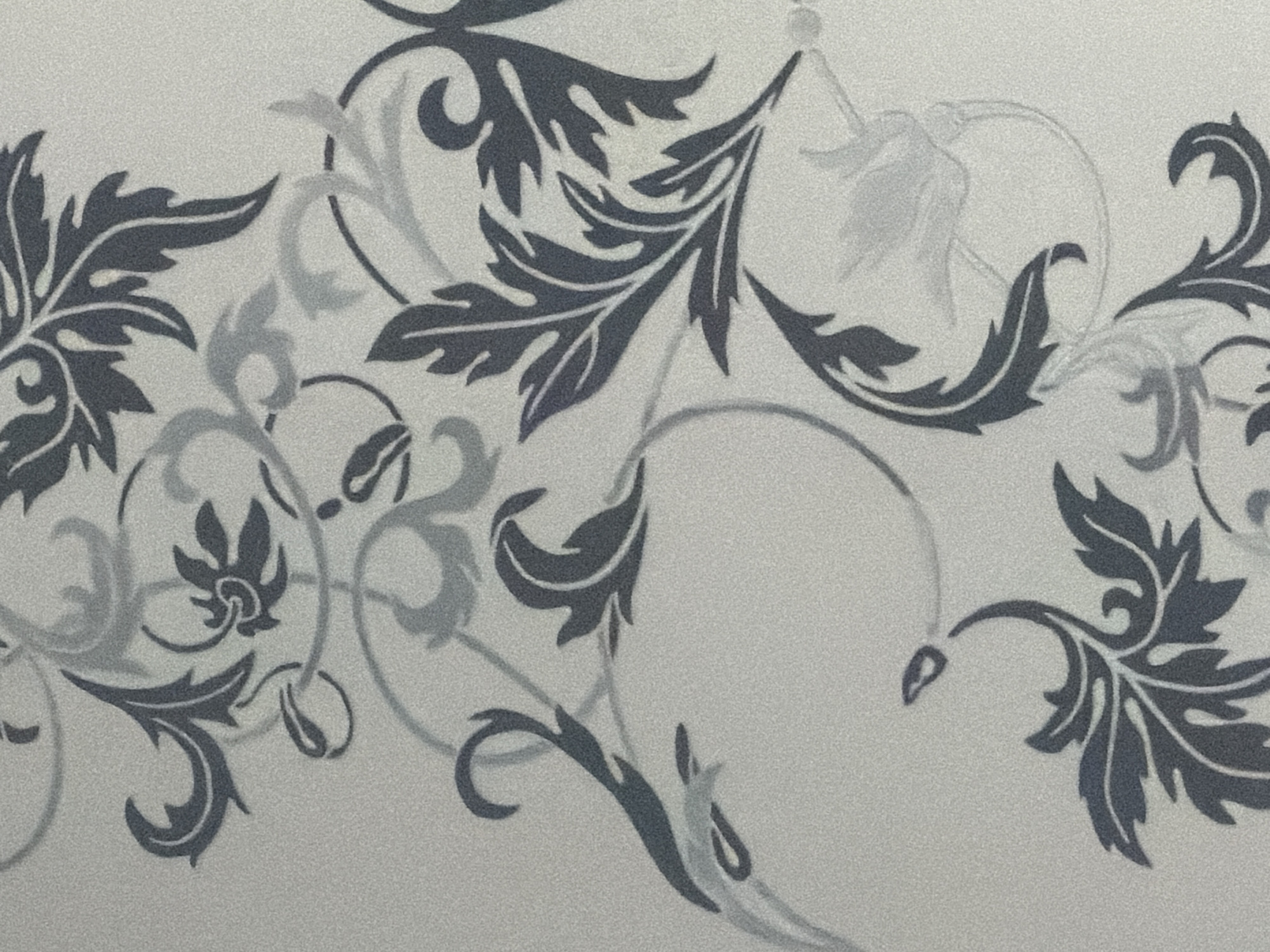 This media file is a close-up, horizontal photograph of a textured, light-colored surface adorned with a repeating, dark gray and lighter gray floral or vine-like pattern. The pattern consists of stylized leaves and swirling tendrils. Some elements of the pattern are solid dark gray, while others appear as lighter gray outlines or faded versions of the darker elements, suggesting either a layering effect or a subtle shading within the design. The surface itself has a fine, granular texture, resembling fabric, wallpaper, or a textured paper. There are no people or discernible objects within the frame, and the activity or event is simply the presentation of this decorative pattern. The lighting is even, and there are no indicators of time of day, weather, or specific location. The image is devoid of any visible text.