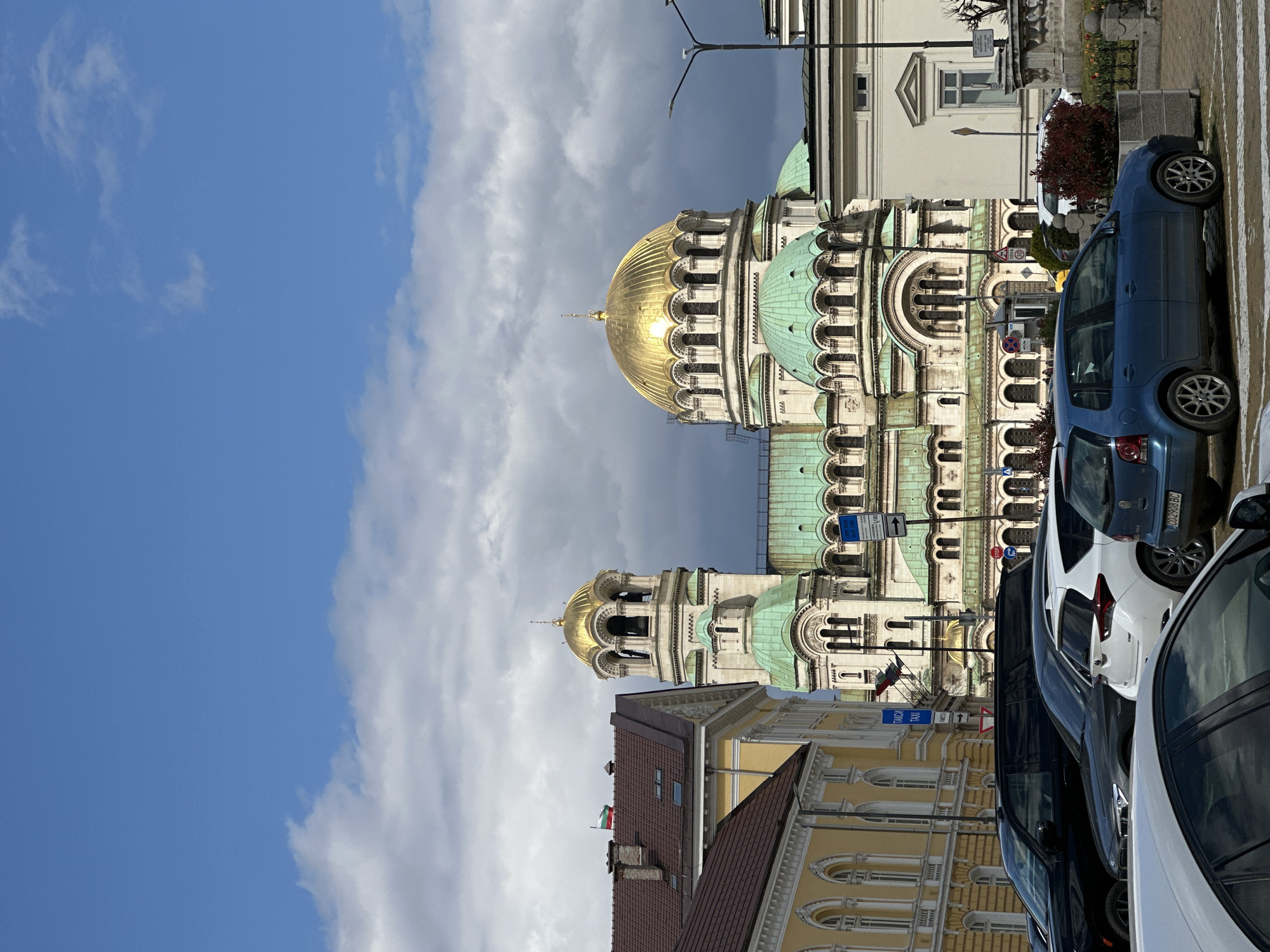 The image shows the St. Alexander Nevsky Cathedral in Sofia, Bulgaria. The cathedral's domes, some gold, are visible against a partly cloudy sky. In the foreground are parked cars of various colors and models. Buildings with architectural details are also visible, including sections with yellow facades. The view is taken at street level.