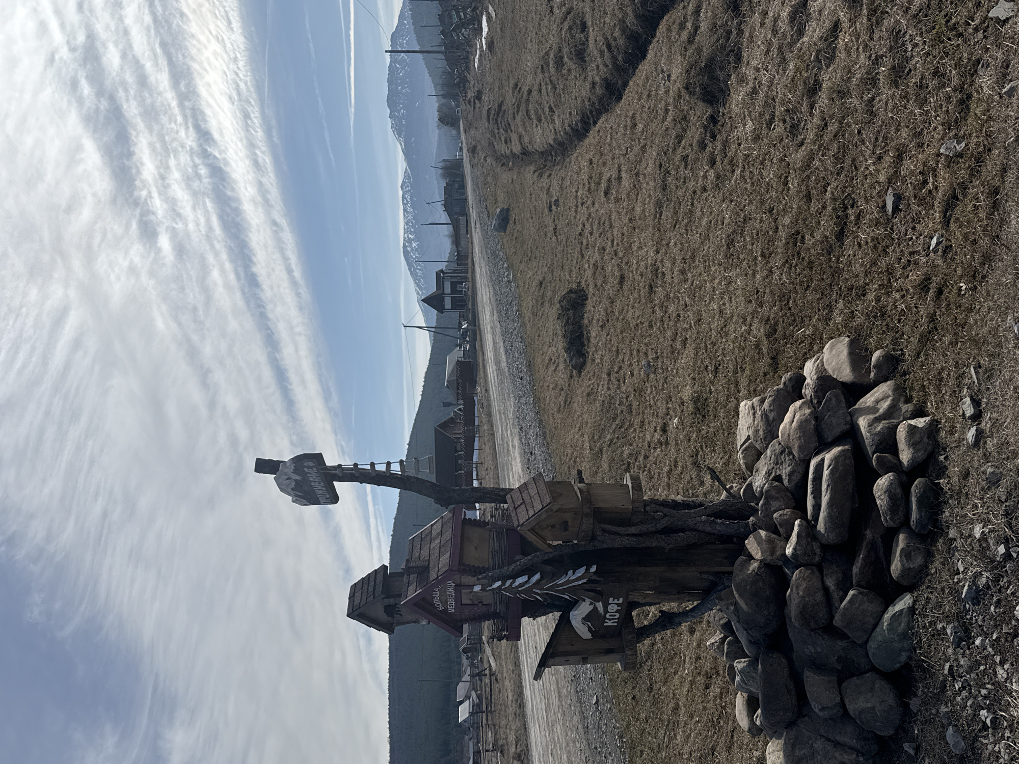The image shows a landscape in Tyulyuk, Russia, featuring a road with buildings and mountains in the background. A wooden, multi-tiered structure sits in the foreground, surrounded by rocks. The structure has signs in Russian, including "проживание" (accommodation), "большая медведица" (Big Dipper), and "кофе" (coffee). The sky is partly cloudy with visible contrails. The surrounding area appears to be dry grass and dirt.