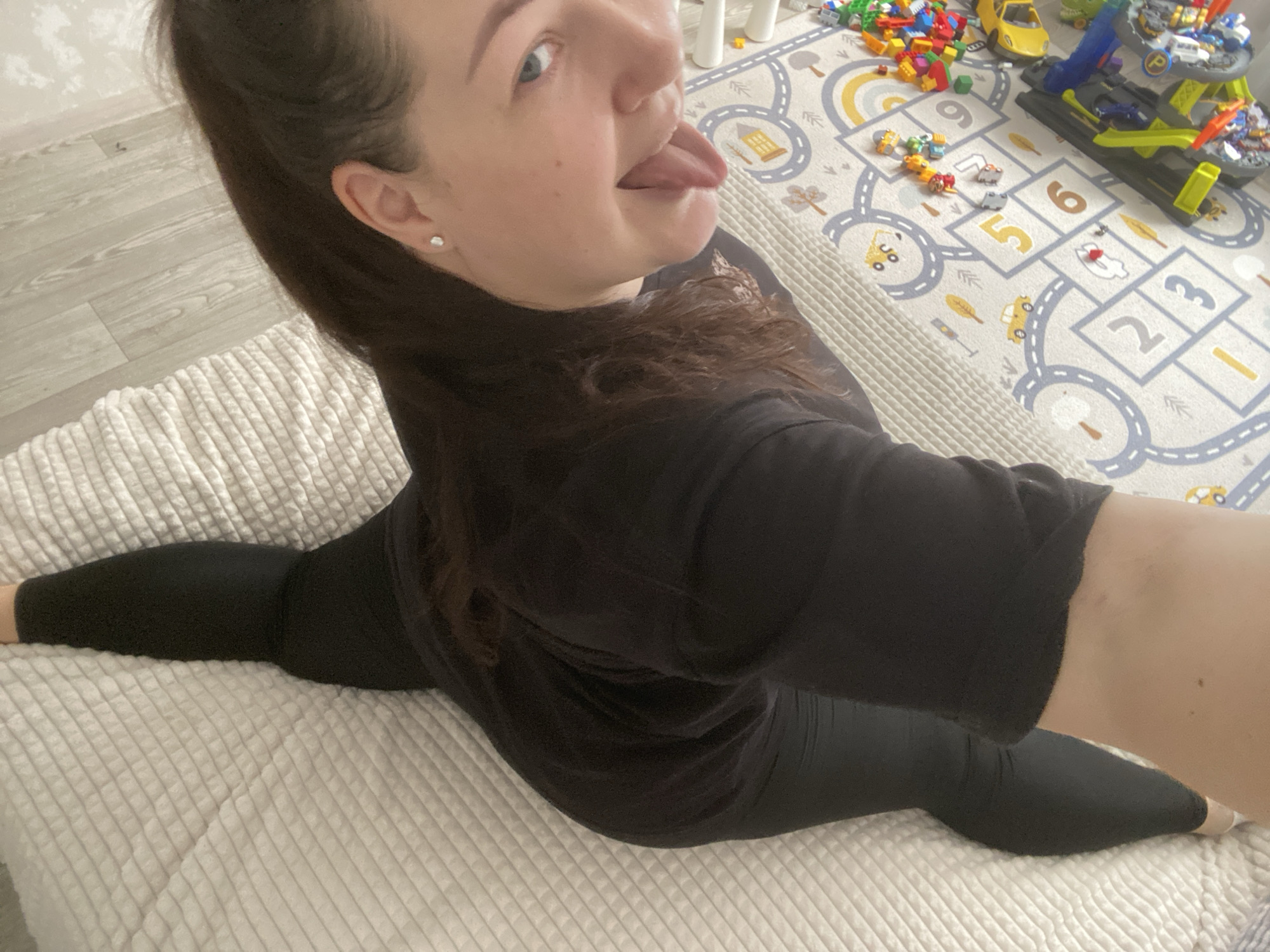 A woman in black leggings and a black top is doing the splits on a white textured surface, possibly a bed. She is looking up at the camera with her tongue out. A colorful children's playmat with numbers and cars is visible in the background, along with a pile of toys. The location is unknown and cannot be determined to be in Sochi, Russia, based on the image.