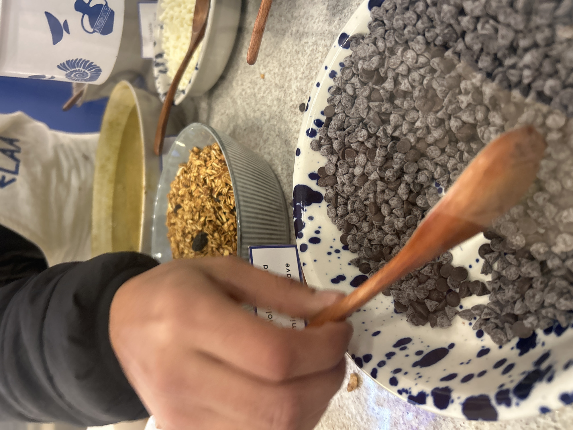 The image shows a person using a wooden spoon to scoop chocolate chips from a large white and blue speckled bowl. The bowl is filled with a variety of chocolate chips. A bowl of granola and a bowl of white shredded cheese are visible in the background. A cup with blue designs is also visible. A tag with text can also be seen by the bowl of granola. The location is not discernible.