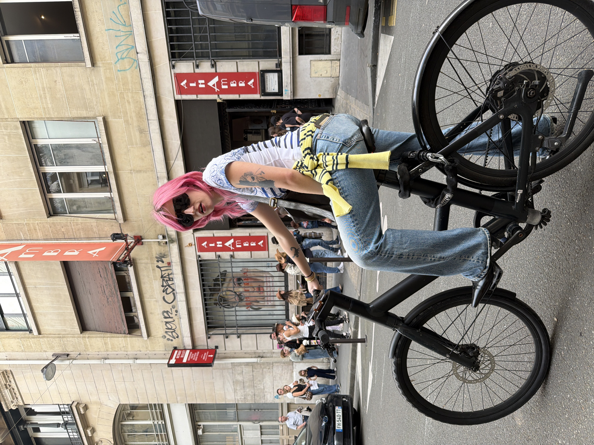 A person with pink hair, wearing sunglasses, a striped shirt, and jeans, is straddling a black bicycle on a city street. The person has a tattoo on their arm and a yellow and black striped garment tied around their waist. The bicycle is locked with a chain. A group of people stand on the sidewalk in the background, along with a black car parked at the curb. Building facades line the street, some with red signs featuring French text, likely business establishments. Graffiti is visible on one of the buildings. A sign for "AU HIBOU" is clearly visible. The location is the city of Paris, France.
