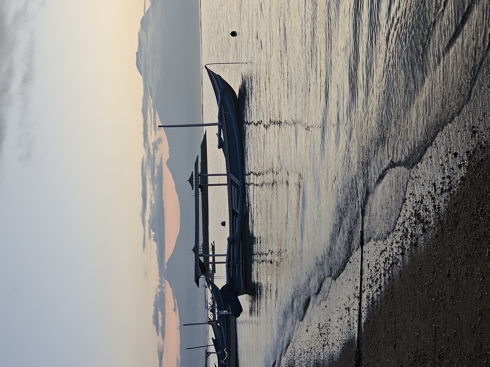 The image shows a scene at sunrise or sunset on a beach in Denpasar, Indonesia. A traditional outrigger boat, is partially submerged in the shallow water near the shore. The boat is likely a "jukung," a type of canoe common in Indonesia. The water reflects the light of the sky, creating a shimmering effect. The beach itself appears to be dark sand, transitioning from wet sand near the water to drier sand further up the shore. In the background, there are landmasses, likely mountains or hills, partially obscured by haze or clouds, with hints of orange and pink hues, indicating a sunrise or sunset. The sky is light grey. The scene is quiet and peaceful.