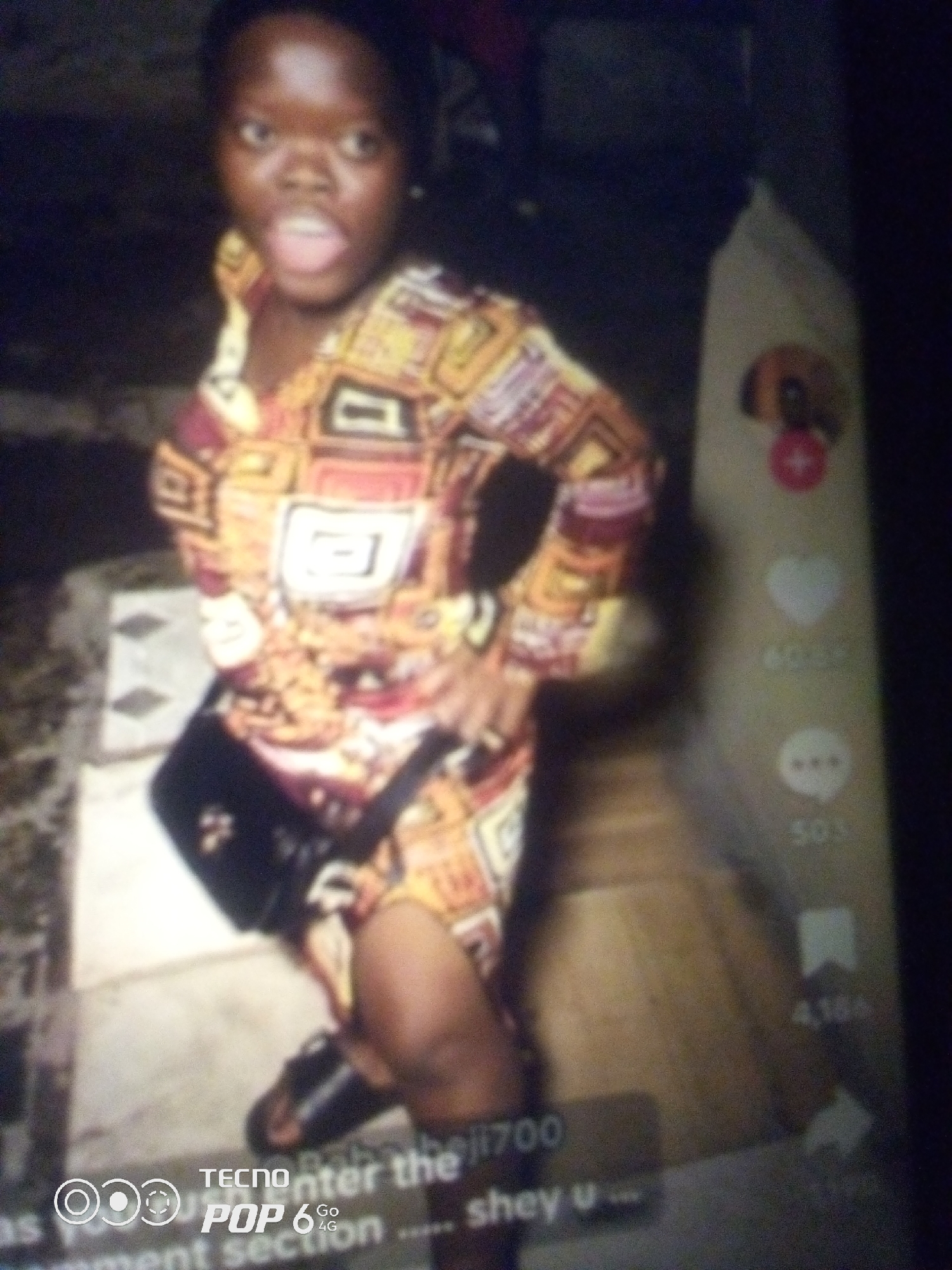 The image shows a young woman with dark skin, wearing a brightly colored, patterned dress and a black crossbody bag. She is standing outdoors, possibly in a courtyard or on steps, with a wall or building behind her. The lighting is dim, suggesting it might be nighttime or an indoor setting with low light. There are some digital interface elements visible, like icons for likes, comments, and shares, along with text that appears to be from a social media platform. The woman is looking at the camera with her mouth slightly open. The lower part of the image displays a watermark with "TECNO POP 6 GO 4G". It is not possible to determine the specific location in the city of Howayi, Nigeria from the visual information provided.
