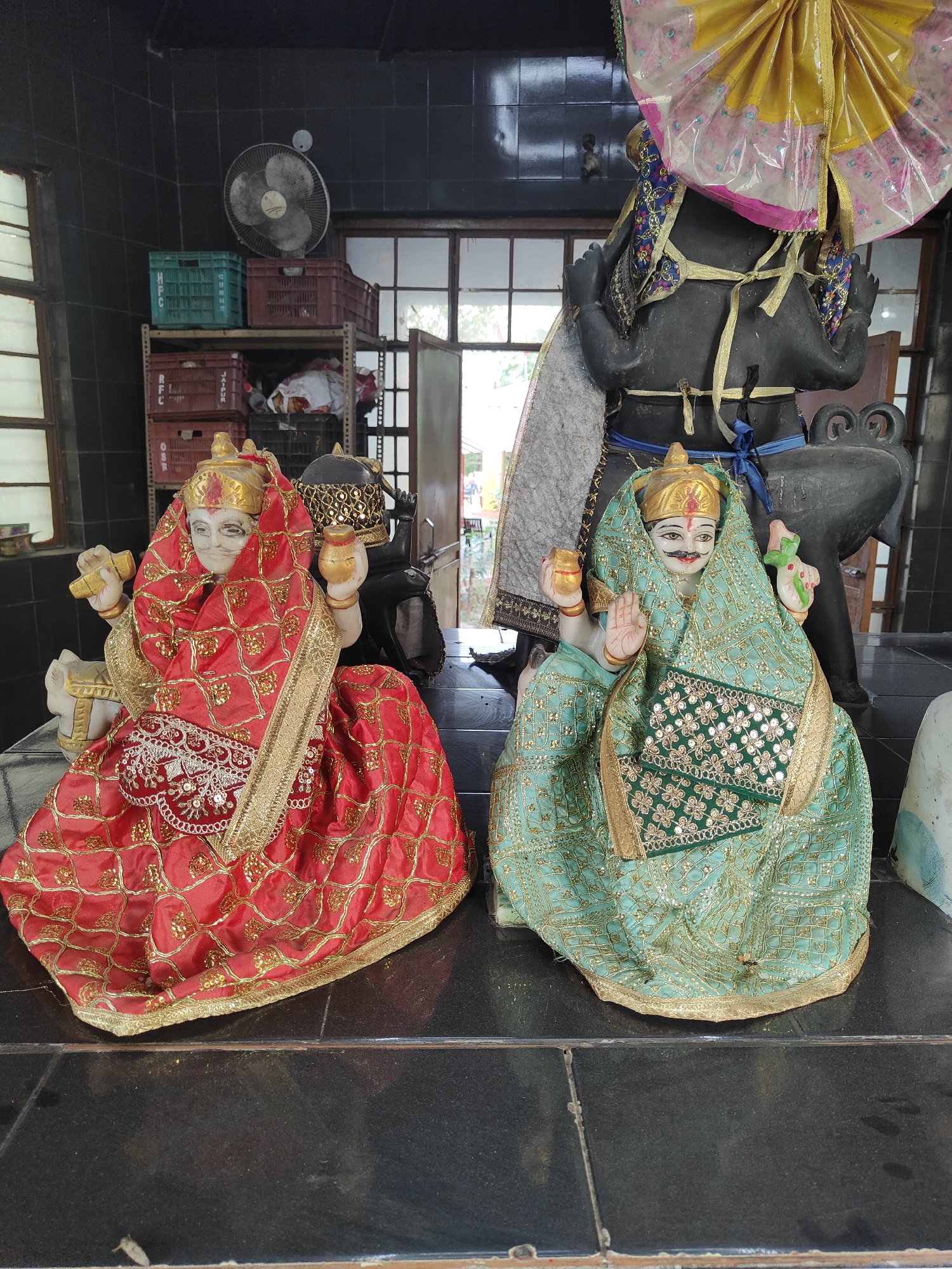 The image captures an indoor scene at a temple in Gurgaon, India. The foreground features two statues of deities, likely part of a religious shrine. The statue on the left is adorned in a vibrant red and gold sari, while the statue on the right wears a green and gold sari. Both statues have distinct facial features and are posed with their hands raised. Behind the statues, a large black idol is partially visible, topped with a colorful umbrella-like headdress. The background reveals shelves stocked with crates and other assorted items, suggesting a storage area within the temple premises. A wall-mounted fan is also visible on the tiled wall in the upper left quadrant of the image. The overall setting appears to be a modest religious space, possibly a community temple.