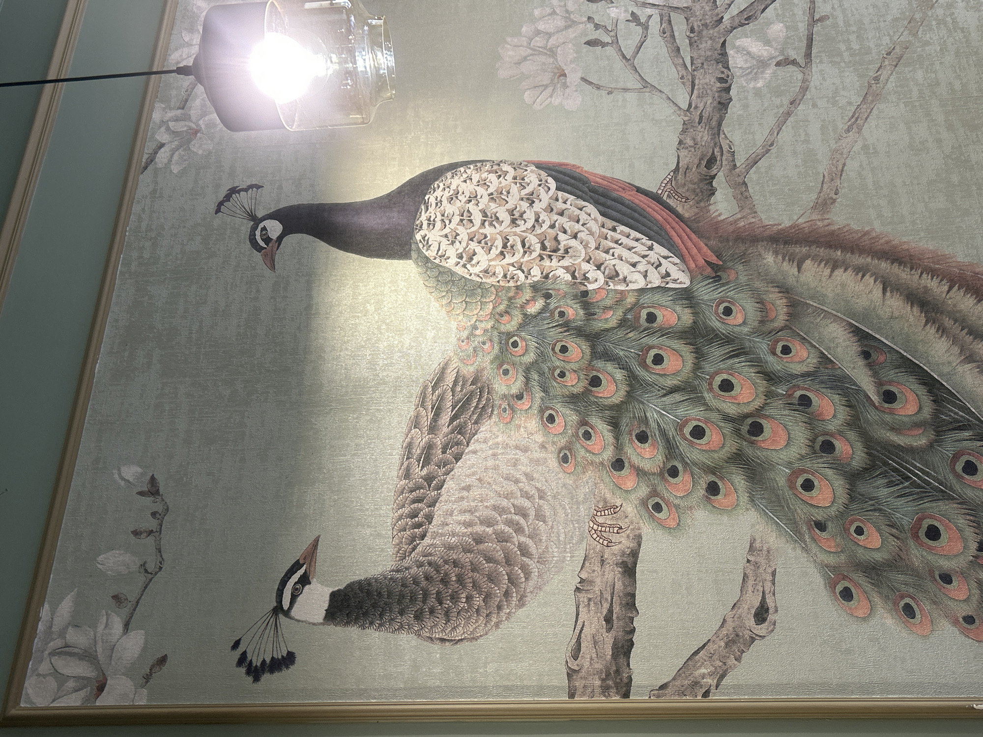 The image displays a framed artwork featuring two peacocks perched on a tree branch against a textured, pale green background. The left peacock is facing left, with its head turned towards the viewer, showcasing its intricate feathering. The right peacock is facing right, with its back to the viewer, and its vibrant, eye-spotted tail feathers are prominently displayed. The artwork also depicts stylized white flowers and branches. A modern light fixture with a clear glass shade and a visible lightbulb hangs from the ceiling in the upper right portion of the image, casting a bright light. The room appears to have a subtle, pale green wall color above the artwork. The artwork is likely located in an indoor establishment in Almaty, Kazakhstan, such as a cafe, restaurant, or home, given the presence of the decorative artwork and the ceiling light fixture.