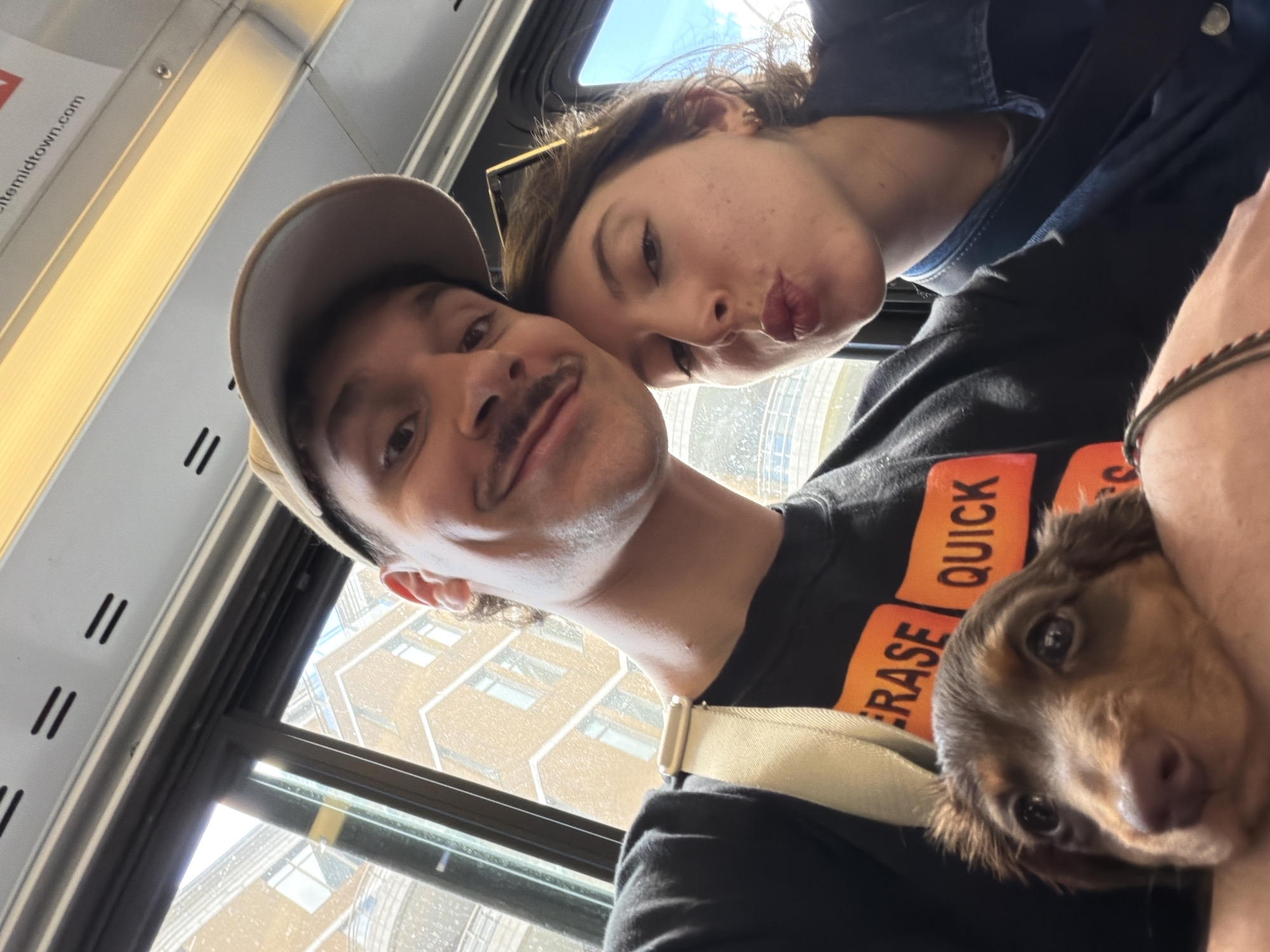 A selfie-style photograph shows a man and woman, and a dog, inside a public transit vehicle. The man, wearing a baseball cap and a black shirt with orange lettering, is in the foreground. The woman is leaning in, kissing his cheek. A small dog is nestled in the man's arms, partially obscured by the clothing. Outside the window, buildings and sky are visible, identifiable as the city of Montreal, Canada due to the architectural style of buildings and overall urban landscape. The transit vehicle has vertical lines and a bright overhead light.