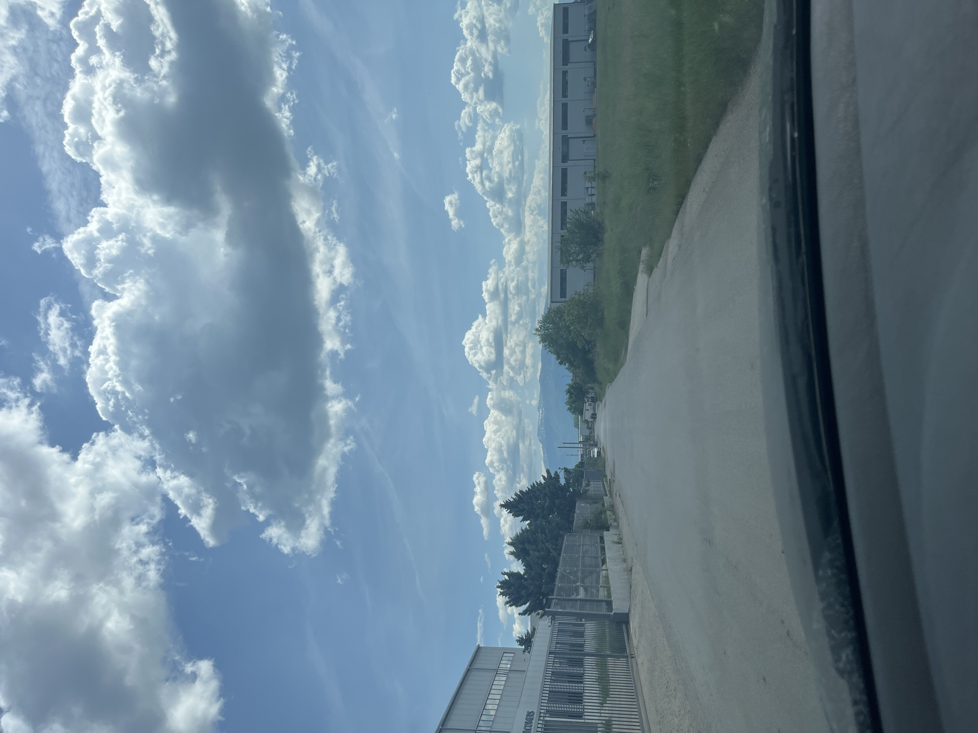 The image depicts a view from a vehicle, potentially a car, traveling on a road in Mirovyane, Bulgaria. The camera is positioned inside the vehicle looking out towards the road and the surroundings. On the right side of the road, there is a grassy area and a building complex. A concrete road is visible, leading into the distance. On the left side of the road, there is a gray or silver industrial building with a tall security fence. Above, there is a bright blue sky with scattered white clouds. Some trees and utility poles are also visible along the road. The environment suggests a rural or industrial outskirts of the city, with a combination of natural and man-made structures.