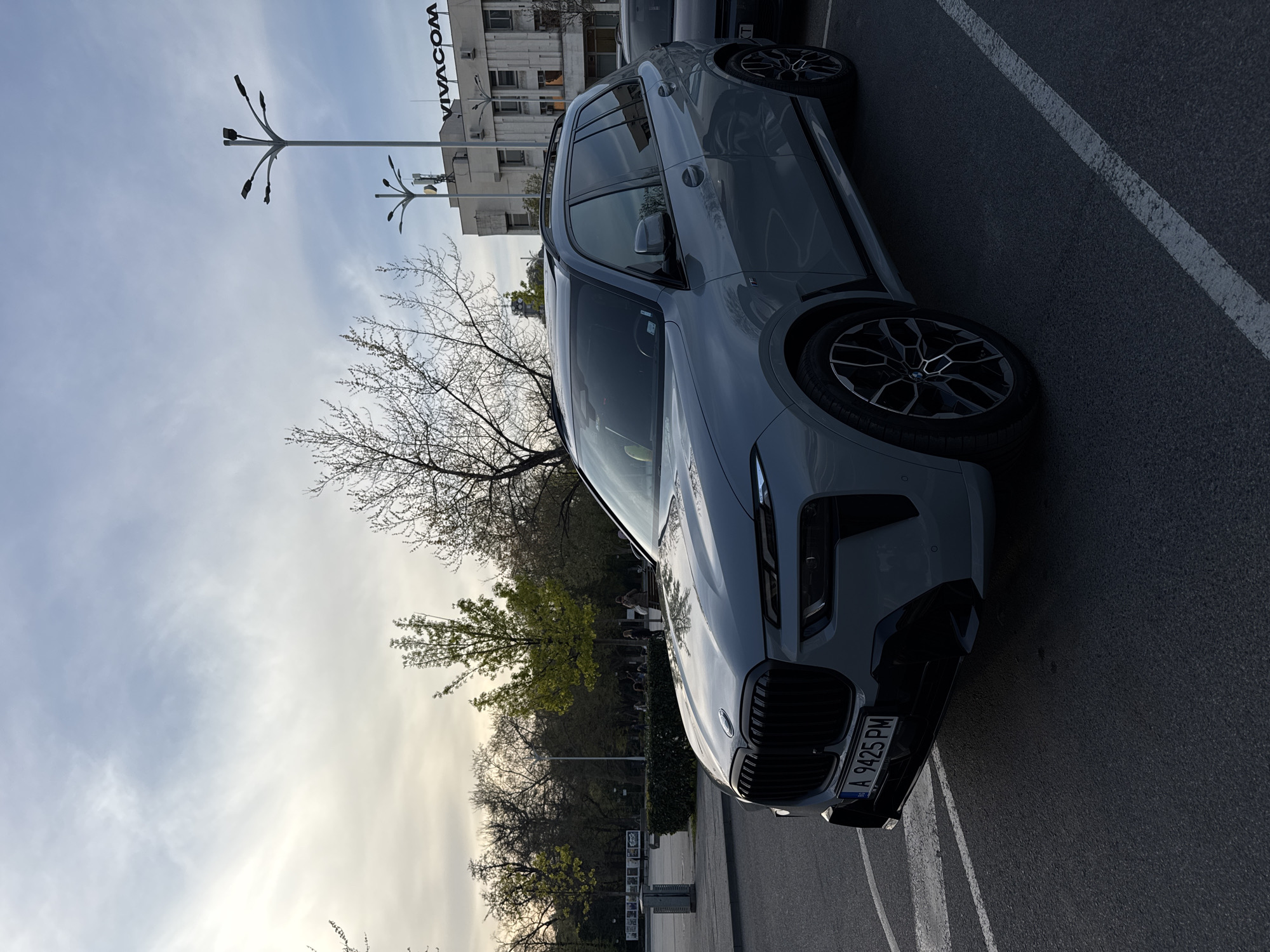 A light gray BMW X5 is parked in an asphalt parking lot. The license plate reads "A 9425 PM." The front of the car is in the foreground. Behind the car is a row of trees. In the background, a light-colored building with the word "VIVACOM" on the side is visible. A streetlight with multiple arms extends upwards on the left. The sky is partly cloudy. The location is Plovdiv, Bulgaria.