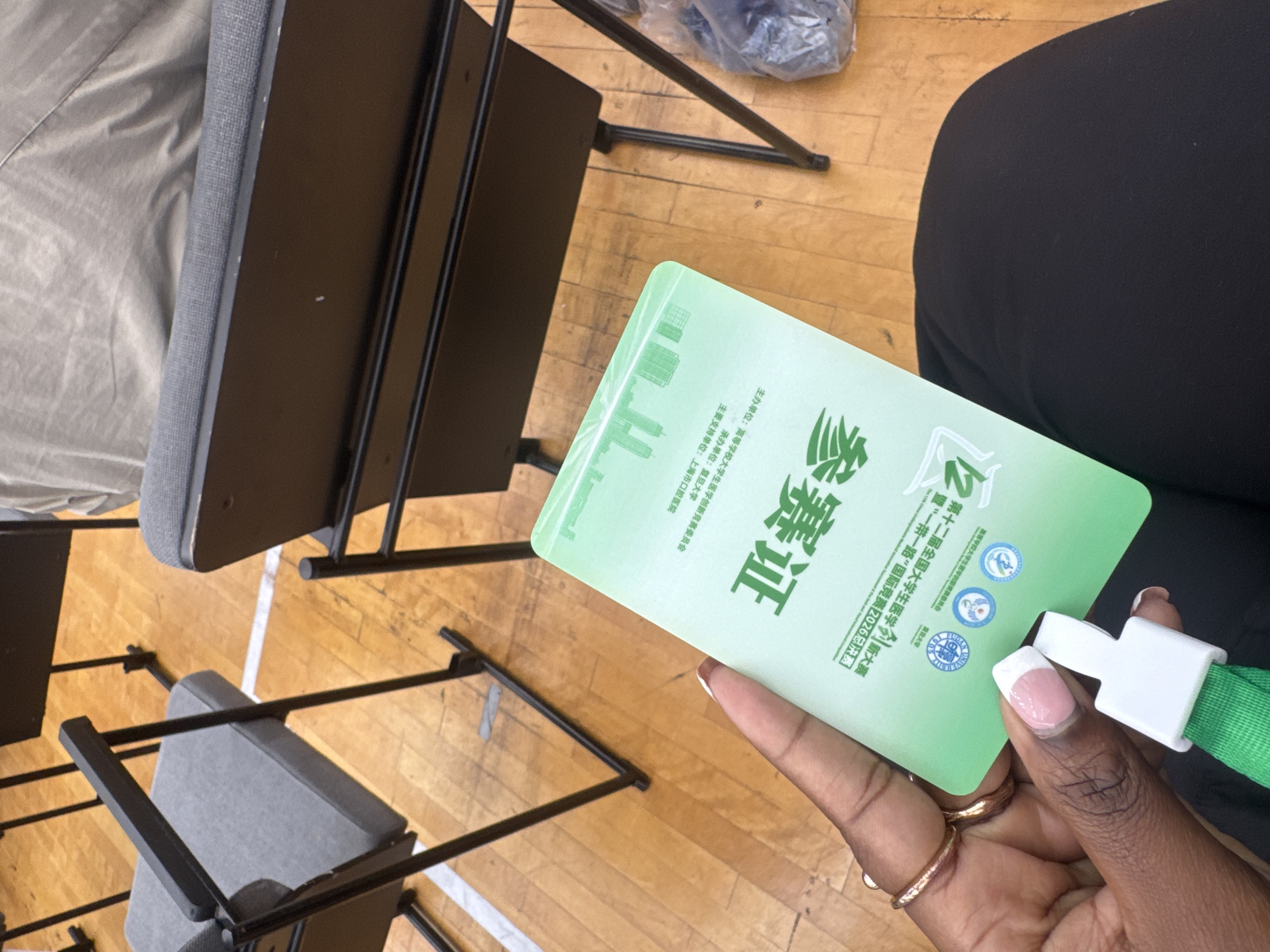 A person holds a green identification badge with Chinese text and logos while seated on a wooden floor with rows of black chairs in the background. The badge appears to be for an event, with text indicating "The 12th 'One Belt One Road' International Student Entrepreneurship Competition 2026". The surrounding environment suggests an indoor venue, possibly a gymnasium or conference hall, with the characteristic polished wooden floor and white line markings seen in such locations in Yangpu District, China. The person's hand, adorned with rings and manicured nails, is visible on the lower left of the image. The chairs have grey cushioned seats and backs, and black metal frames. Some plastic-wrapped items are visible in the background on the right.