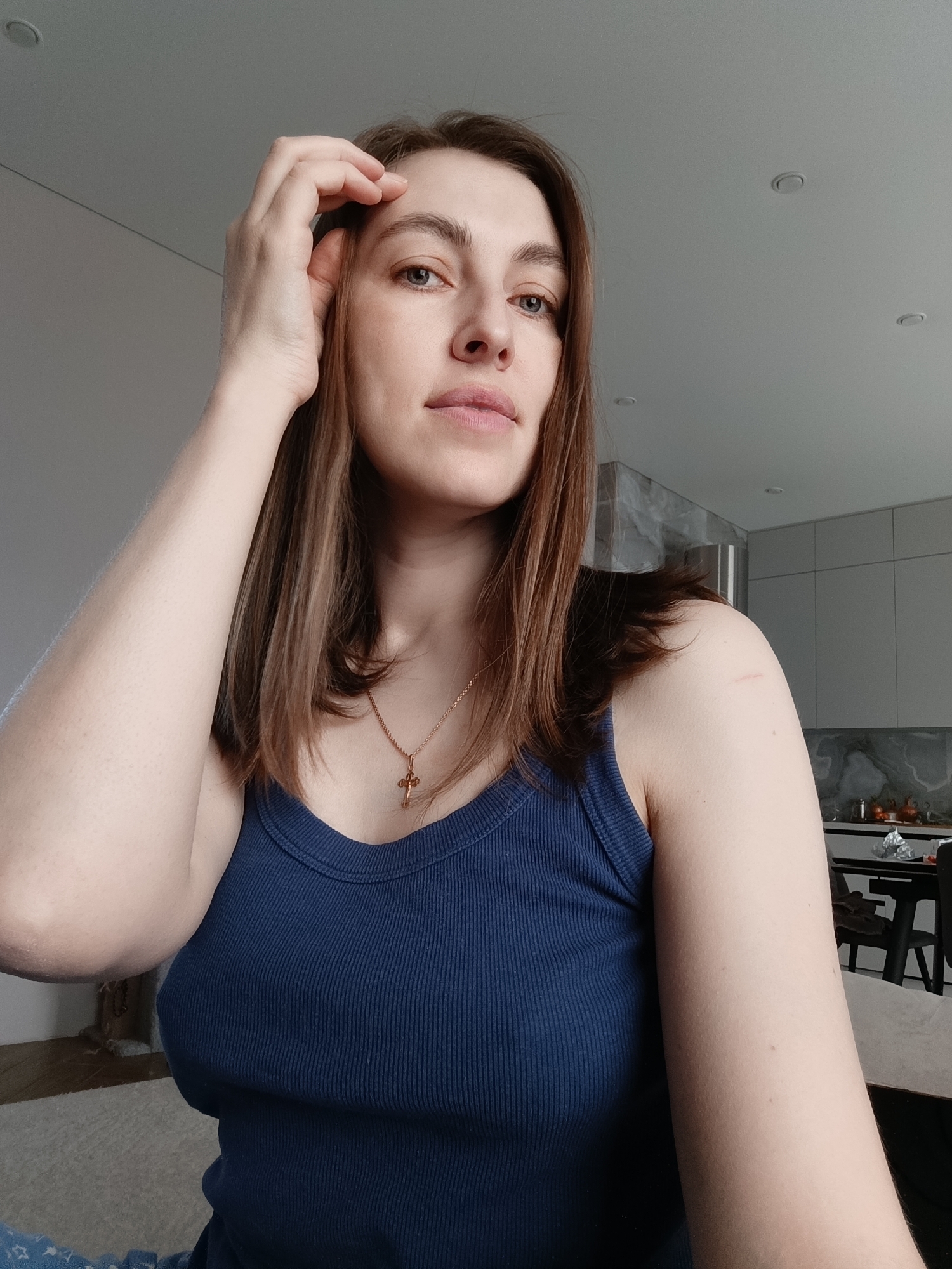 A woman is depicted in a close-up, head-and-torso shot, looking directly at the camera with a neutral to slightly contemplative expression. She has shoulder-length brown hair, fair skin, and light-colored eyes. Her right hand is raised, touching her forehead near her hairline. She is wearing a dark blue, ribbed tank top and a gold chain necklace with a small crucifix pendant. A faint scar or mark is visible on her left shoulder.
The setting is indoors, featuring a modern and minimalist aesthetic. The background consists of light-colored, possibly white or light grey, walls and a white ceiling with recessed spotlights. To her right, a partial view of a kitchen or dining area is visible, featuring sleek grey cabinets and a dark table with chairs. The lighting is bright and even, suggesting it is daytime or a well-lit interior. There is no visible text in the image. The image was taken in Chaikovsky, Russia.