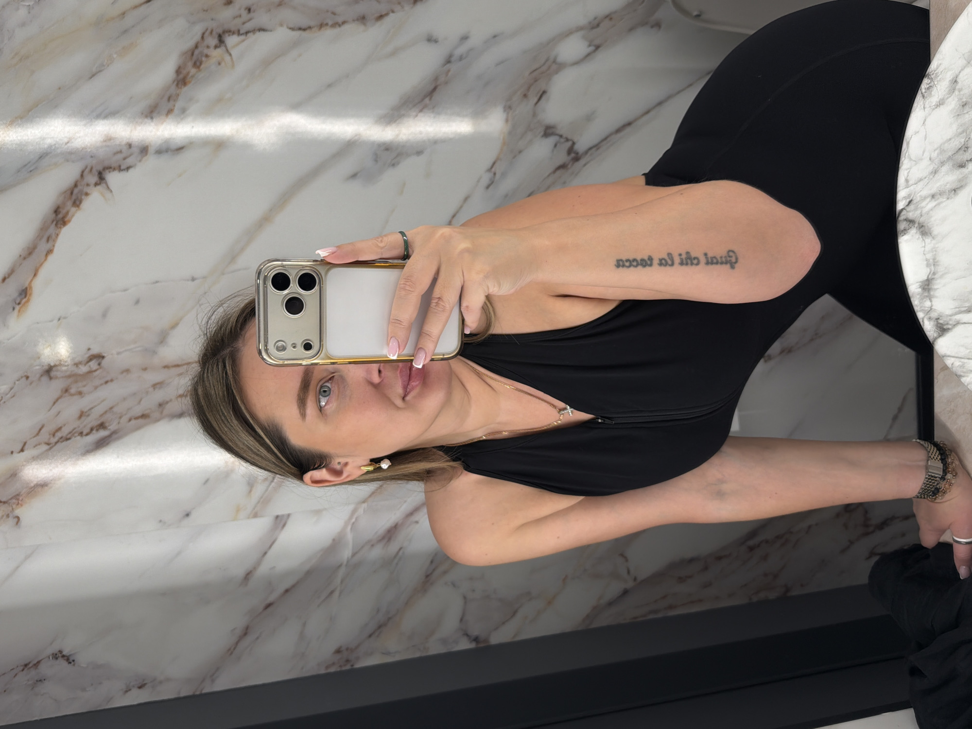 A woman takes a selfie in a bathroom with marble-patterned walls, seemingly in Moscow, Russia. She is wearing a black outfit, holds an iPhone in her left hand, and has a tattoo on her right forearm. She also wears a gold necklace and pearl earrings. The floor is bordered with a dark gray trim. Part of a white marble countertop is visible to the right.