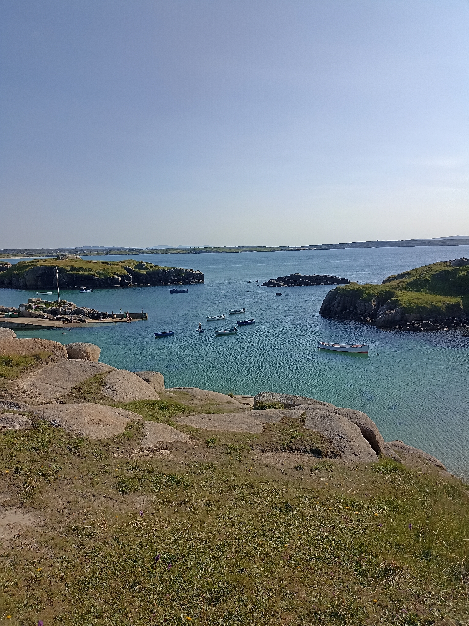 This is a high-angle, long shot photograph of a coastal scene in Carnboy, Ireland. The image features a body of water with several small boats, both moored and adrift. The water is a clear, turquoise color, with visible ripples. Rocky islands and a small pier are present in the water, with some people visible on the pier and in the water. The foreground is dominated by large, weathered rocks and grassy terrain with scattered wildflowers. The background consists of more islands and a hazy horizon under a clear blue sky. The overall scene depicts a tranquil, natural landscape.