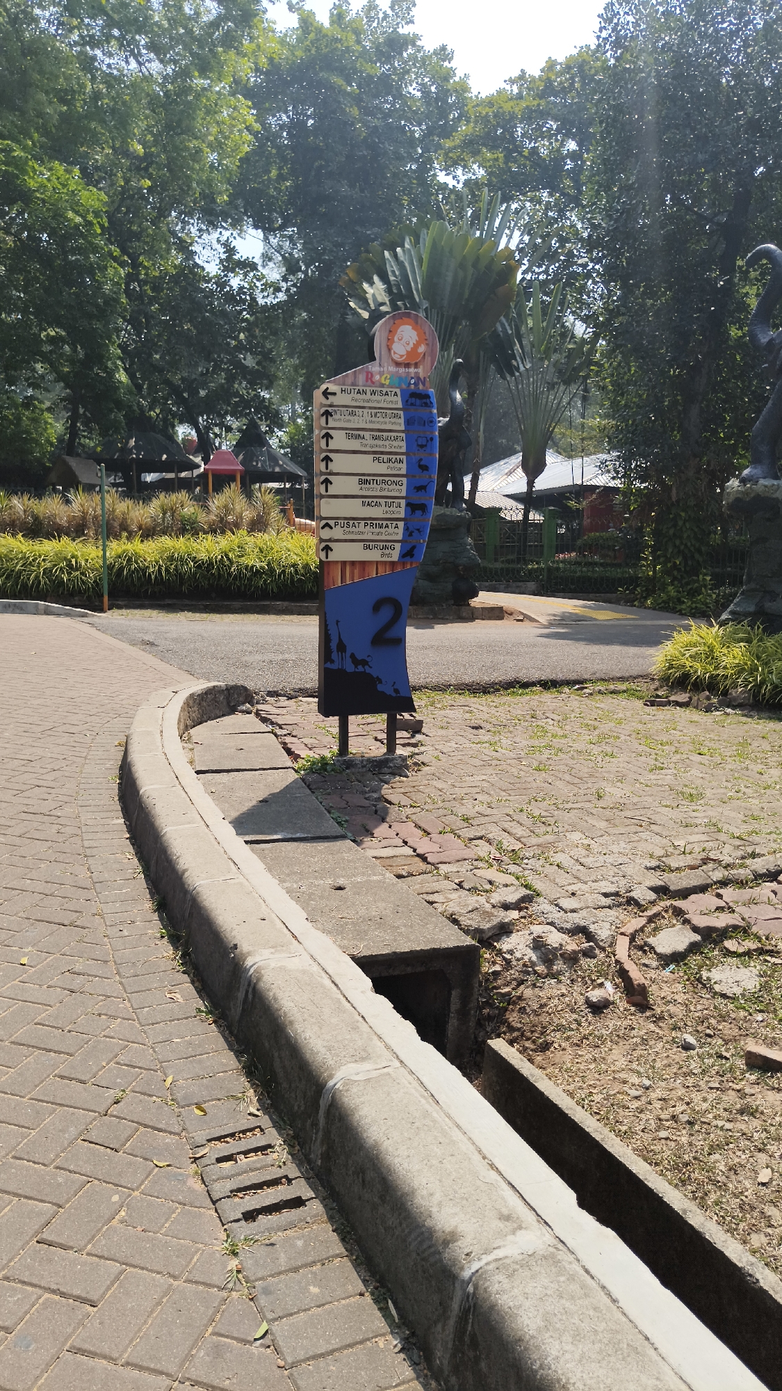 In South Jakarta, Indonesia, a directional signpost stands tall amidst a lush green environment. The signpost, adorned with various animal silhouettes and text, points towards different attractions. One arrow indicates "Hutan Wisata" (Tourist Forest), another directs towards "Pintu Utara, 1 & 2 Motor Utara" (North Gate, 1 & 2 North Motor), and another points to "Terminal, Transjakarta" (Transjakarta Terminal). Additionally, the sign highlights "Pelikan" (Pelican), "Binturong" (Bearcat), "Macan Tutul" (Leopard), "Pusat Primata" (Primate Center), and "Burung" (Bird). The number "2" is prominently displayed on the sign. Surrounding the signpost, the ground is paved with interlocking bricks and concrete. A drainage ditch runs alongside the paved area. In the background, trees and foliage create a natural canopy, with glimpses of thatched-roof structures and playground equipment. The scene is bathed in sunlight, suggesting a clear day.