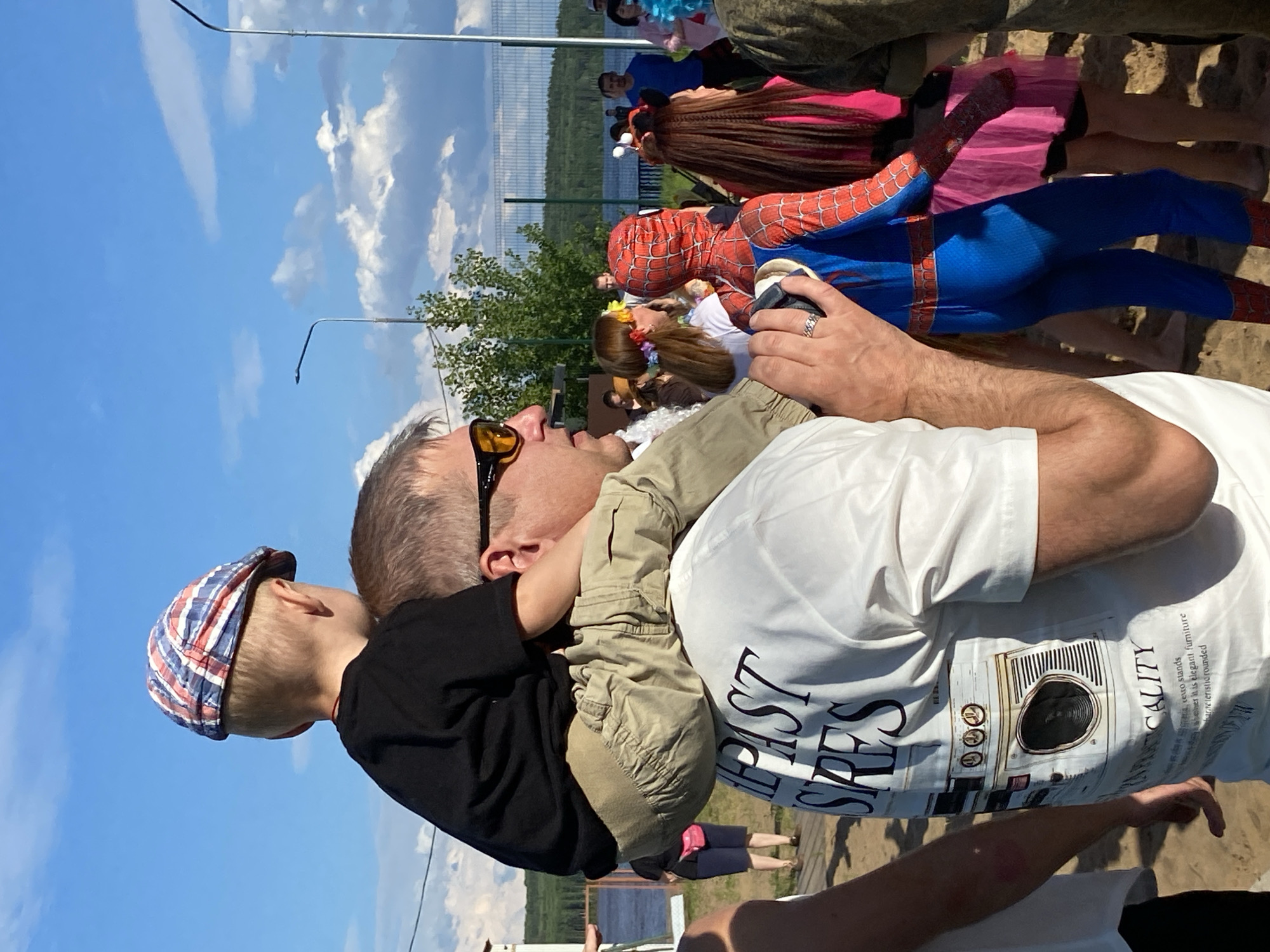 A man carries a young boy on his shoulders in an outdoor setting. The boy wears a black t-shirt and khaki pants, and a red, white, and blue checkered flat cap. The man has on sunglasses and a white t-shirt with a graphic print. In the background, other people are visible, including one person dressed as Spider-Man and another in a pink skirt. The environment appears to be a sandy area near a body of water, possibly a lake or river, with trees and a fence visible in the distance. The sky is blue with scattered clouds, suggesting it is daytime. The context implies a community event or gathering, possibly in Chaikovsky, Russia, given the potential for such events near water bodies in the region during warmer months.
