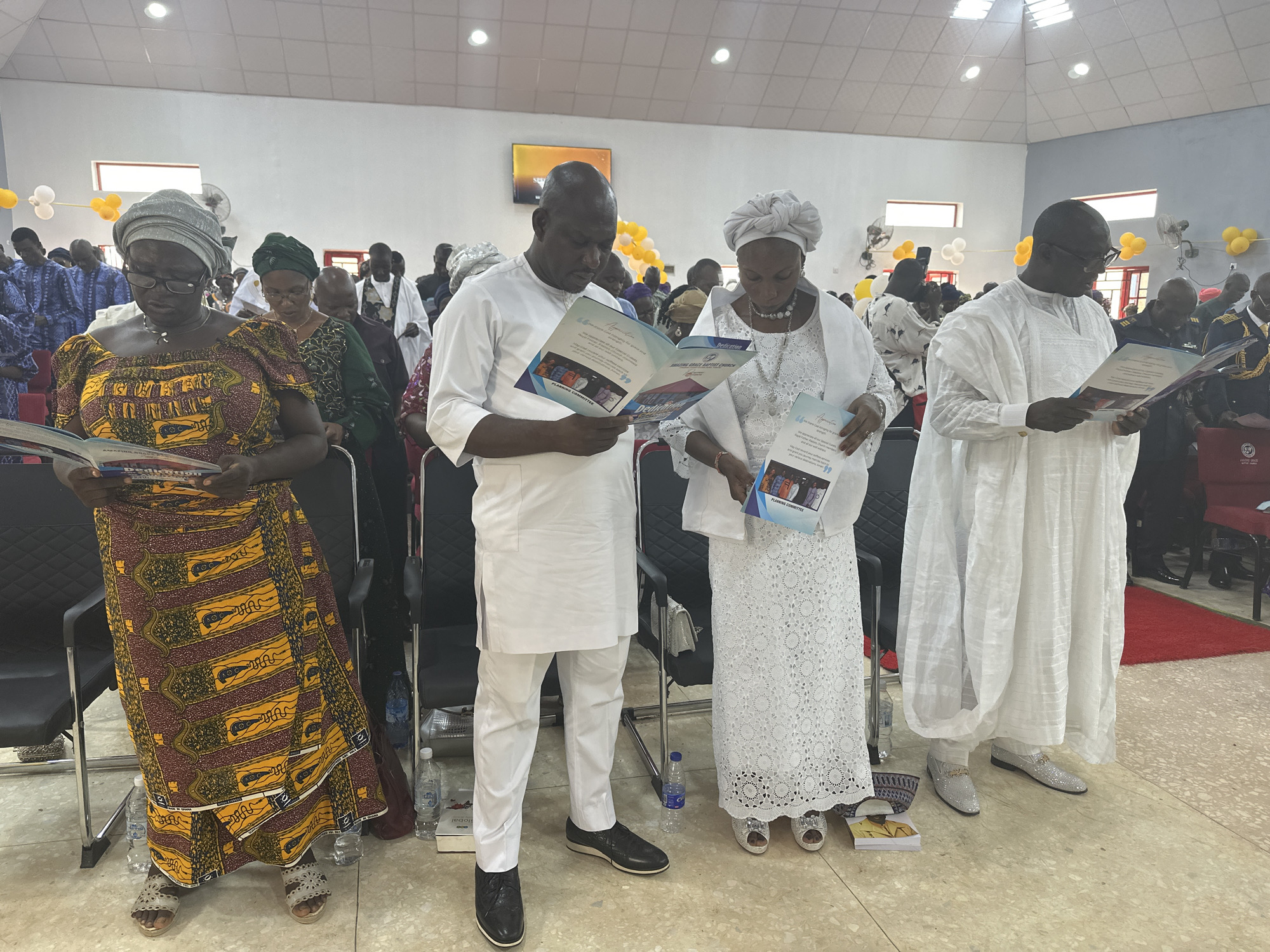 The image depicts a gathering of people inside a building in Goni Gara, Nigeria. Four individuals are standing prominently in the foreground, each holding and reading booklets.
From left to right:
1. A woman wearing a patterned brown and yellow dress with a gray head wrap.
2. A man in a white outfit, reading a booklet with the text "Dedication" and "Planning Committee" visible.
3. A woman in a white lace dress and head wrap, also reading a booklet with the same text.
4. A man in a white outfit with glasses, reading a booklet.
Behind them, a larger group of people is visible, some wearing traditional clothing. The room is brightly lit with a tiled floor. Decorations include balloons and what appears to be a screen with an image or video displayed. Water bottles and books are on the floor near the front group. The overall impression is of a formal gathering or event.