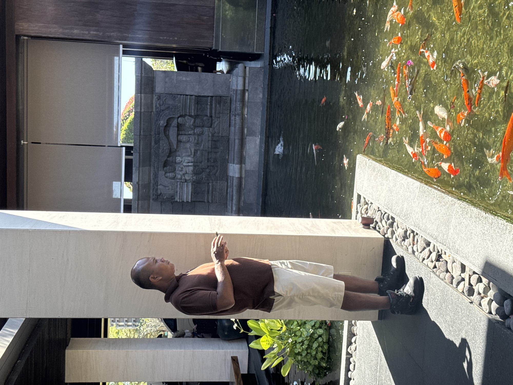 The image depicts a man in Kuta Selatan, Indonesia, standing next to a water feature. He is bald, wearing a brown polo shirt and beige shorts, with dark-colored shoes. He appears to be looking at something in the water, possibly fish, and has his hands clasped together. The water feature is filled with koi fish, with black pebbles lining the edge. There's a grey structure with carved details behind the man, and greenery is visible at the base of the structure. The location is likely outdoors, possibly a resort or hotel.