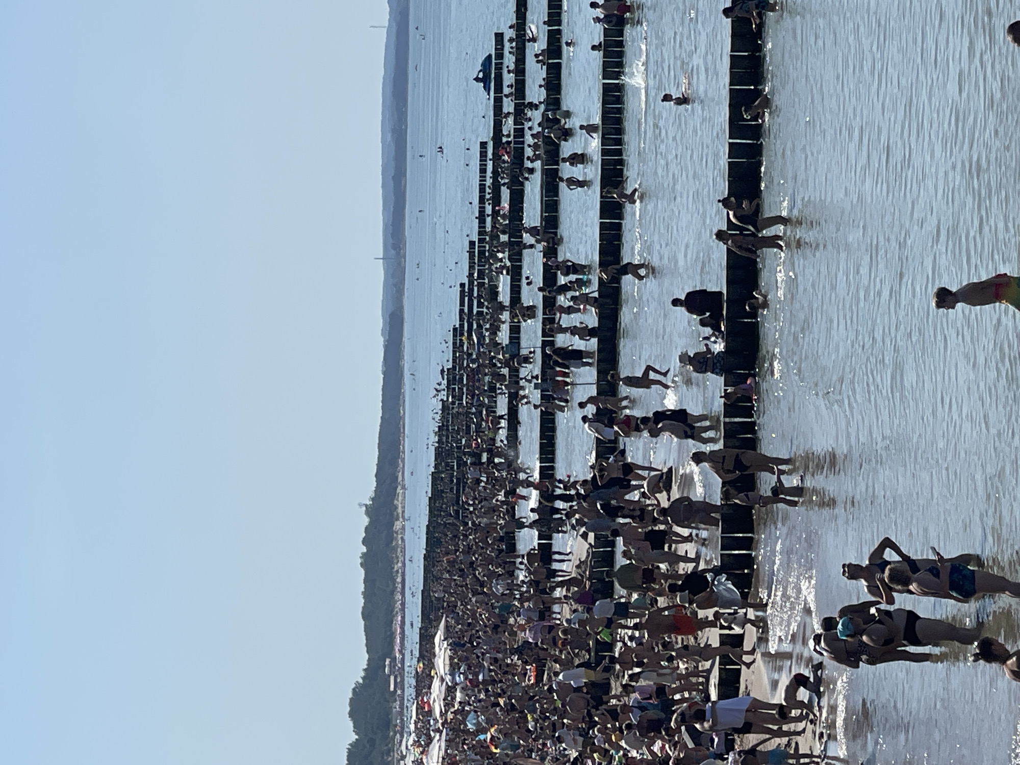 A long, crowded beach in Zelenogradsk, Russia, on a sunny day. A multitude of people are on the sand and in the shallow water. Many are wearing swimwear. Some are walking along the shore, others are wading or swimming. There are rows of wooden breakwaters extending into the sea, creating shallow pools. Beach umbrellas are visible in the background, suggesting a populated tourist area. The sea stretches to the horizon under a clear blue sky. A jet ski is visible in the water in the middle distance.