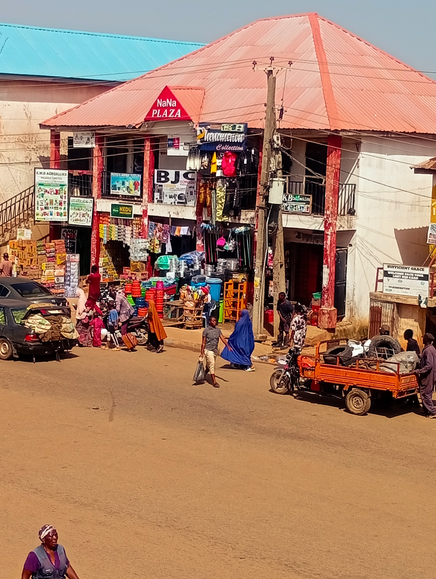 A street scene in Bukuru, Nigeria, shows a busy marketplace in front of a multi-story building. The building has a reddish-orange corrugated metal roof and white walls. A prominent red sign on the front of the building reads "NaNa PLAZA". Various shops are visible on the ground floor, displaying goods such as agricultural products, clothing, and household items.
Several people are interacting in the street. A group of individuals, including children, are gathered around a motorcycle and a car loaded with goods. Some are carrying bags, and one person is pulling a suitcase. Another person in a blue abaya walks across the street. Further down the road, a man walks with shopping bags, and a few men are near an orange three-wheeled vehicle loaded with tires and other items. A woman in a purple top and headscarf is at the bottom left of the frame, looking towards the left.
The street is paved and appears to be a typical urban environment. A utility pole with wires stands prominently in the foreground, with a transformer box attached. The sky is clear and blue. The overall impression is of a lively commercial hub with ongoing activity.